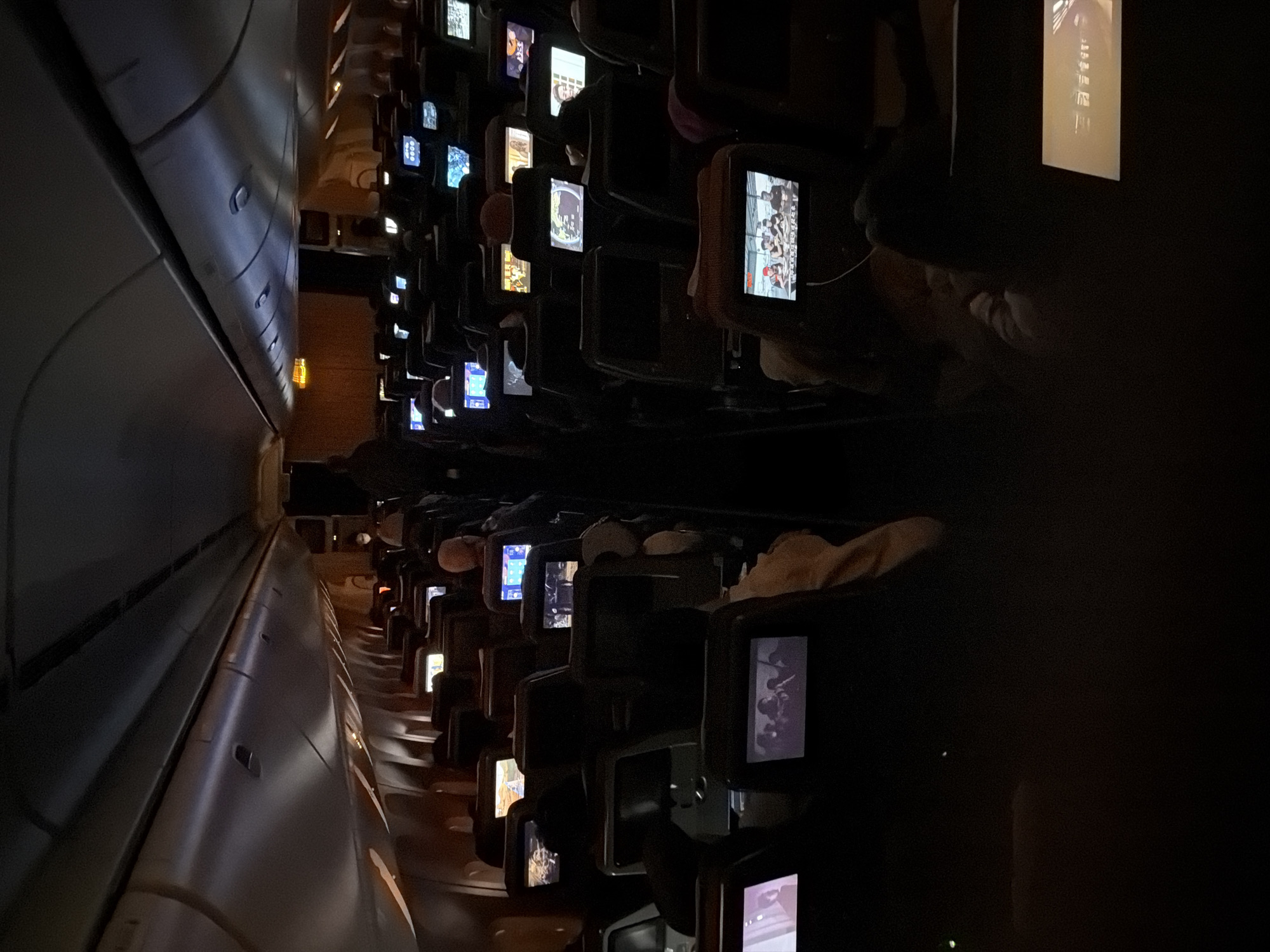 The image shows the interior of an airplane cabin during flight. Rows of seats stretch into the distance on both sides of the aisle, and each seat has an individual entertainment screen displaying various content. Passengers are visible in their seats, some engaged with the screens, others resting. The cabin lighting is dimmed, creating a low-light environment. It's impossible to determine the city of Los Angeles from the image.