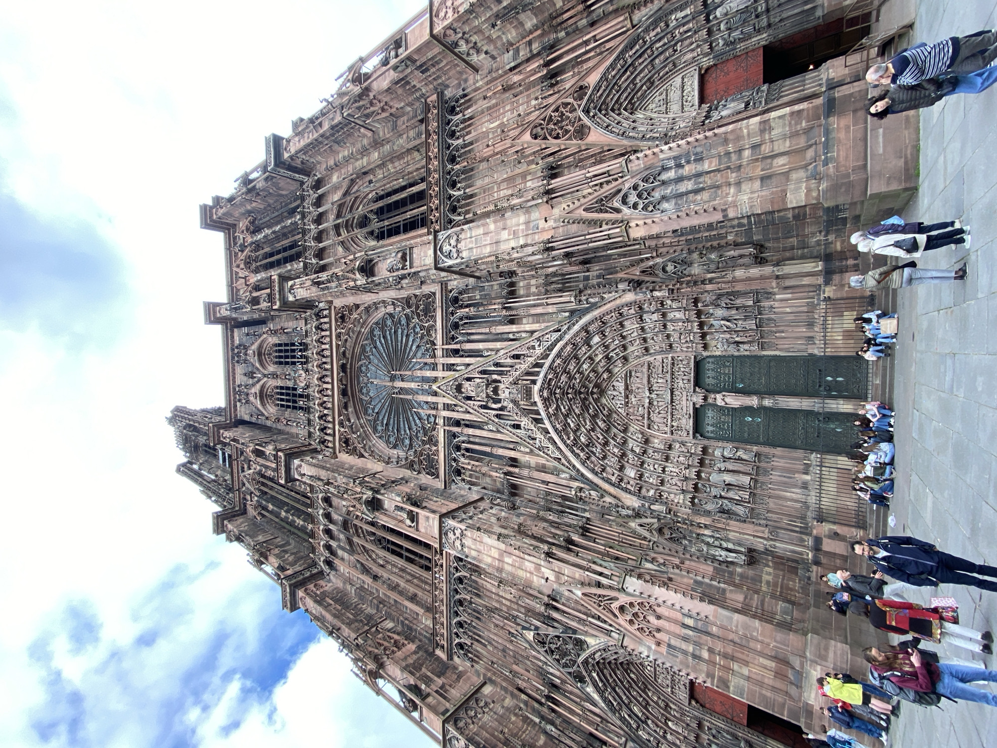 The image depicts the Strasbourg Cathedral in Strasbourg, France. The cathedral's facade is visible, exhibiting its intricate Gothic architecture. A group of people, appearing to be tourists, is present near the base of the cathedral, some standing and others seated. The sky is visible in the background with some clouds.