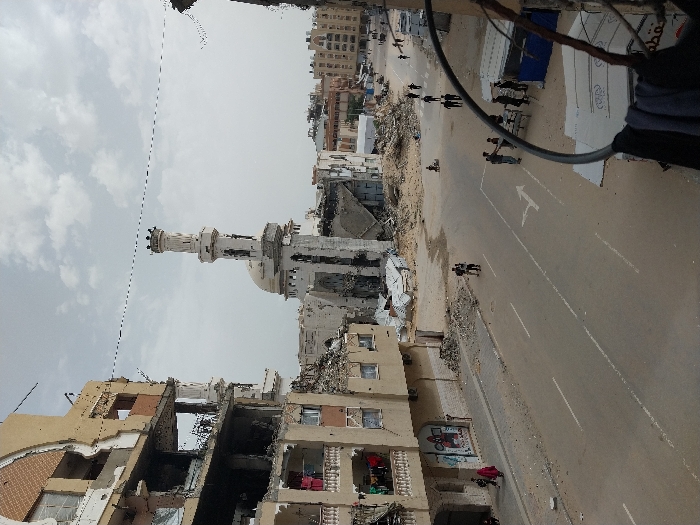 The image captures a wide street in Khan Yunis, Palestinian Territory, showcasing extensive urban destruction under an overcast sky. In the mid-ground, a heavily damaged mosque dominates the center, featuring a partially intact minaret with an ornate top and a damaged dome, surrounded by large piles of rubble from its collapsed structure. To the left, a multi-story building stands severely damaged, with large sections of its facade missing, exposing interior rooms, broken windows, and debris on its balconies. Laundry hangs from a lower balcony of this building, and a poster is visible at street level.
The asphalt road, marked with white lines including a double solid line and a turn arrow, runs through the center of the frame. Debris lines the edges of the road. Numerous individuals are present on the street and sidewalks. Two people walk on the left sidewalk near the damaged building. In the mid-ground, a small figure is visible near the mosque rubble in the street. Further down the right side of the street, a group of people gathers around makeshift structures, possibly stalls. Additional individuals are scattered walking along the road and sidewalks further into the background. In the far background, additional damaged and residential buildings are visible. Overhead, power lines or communication cables traverse the sky. A blurred circular metal object with dark clothing draped over it is prominent in the immediate bottom right foreground.