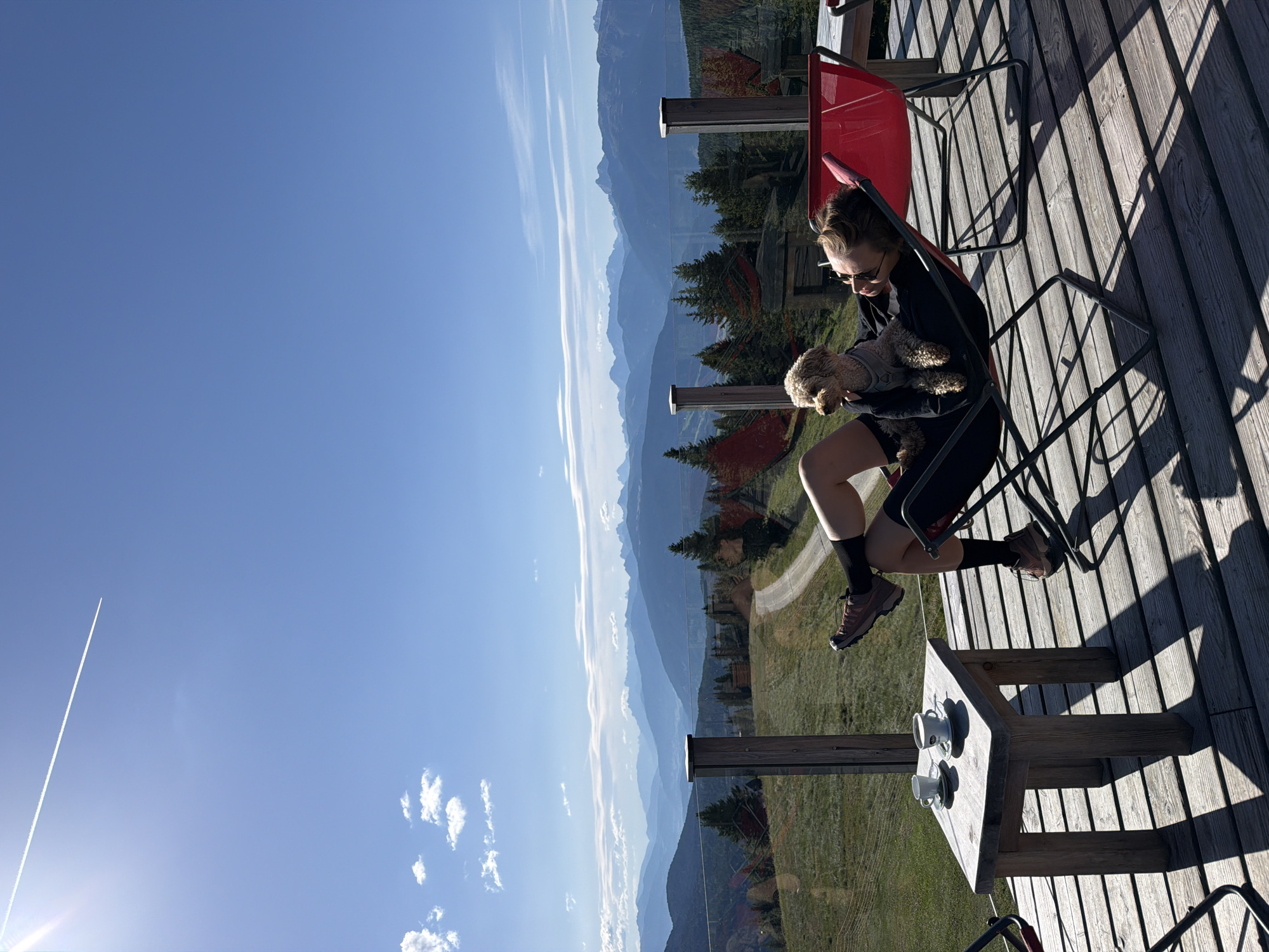 In Mühlbach - Rio di Pusteria, Italy, on a wooden deck overlooking a mountain landscape, a person reclines in a red chair with a small, light-colored dog on their lap. The person is wearing dark clothing and sunglasses. A small wooden table next to the chair holds two cups of coffee. The background features rolling green hills, scattered trees, and distant mountain ranges under a bright blue sky with a contrail from an airplane.