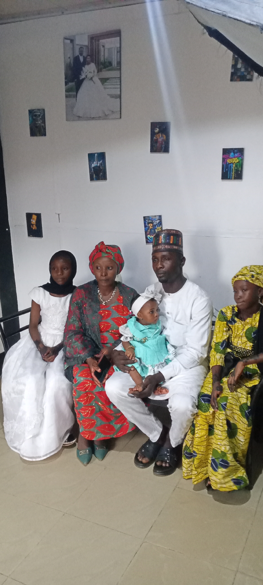 A family of five is posing for a photo in front of a wall decorated with artwork. The adults and children are wearing traditional clothing, the father in a white outfit with a patterned cap, the mother in a red and green patterned dress, and the daughters in a white dress and a yellow patterned dress respectively. The youngest child, a baby, is being held by the father and is wearing a light blue dress with a white headband. The location appears to be indoors, possibly a home, in the city of Unknown, Unknown.