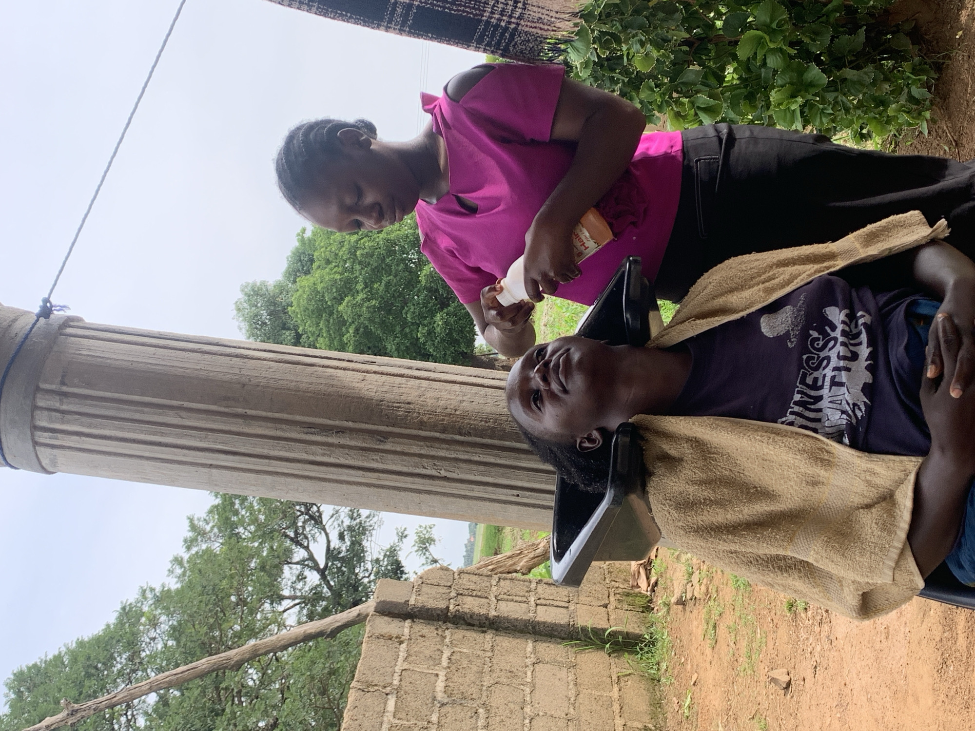 An overhead shot shows two women performing a hair washing and conditioning service outdoors in Agban, Nigeria. A woman sits in a black chair with a tan towel draped over her shoulders, her head tilted back into a black basin. Her hair is dark and braided. The second woman, standing to her right, wears a bright pink short-sleeved top and black pants. She is holding a white bottle with an orange label and is in the process of applying product to the seated woman's hair. The background features a large, fluted concrete pillar, a brick wall, lush green trees, and a cloudy sky. The ground is a mix of dirt and sparse grass.