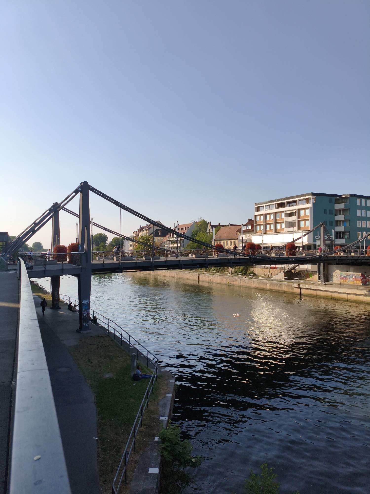 A pedestrian bridge spans a calm river in Bamberg, Germany, under a clear blue sky. The bridge features dark grey metal supports and a wide walkway lined with planters containing red flowers. On the bridge, people are seen walking and cycling. Along the riverbank on the left, a paved walkway is visible with a few individuals walking and sitting. On the right bank, buildings of varying architectural styles line the river, including a prominent modern building with a turquoise facade. The river's surface reflects the sunlight, creating shimmering patterns. The overall scene depicts a peaceful urban environment during daylight hours.