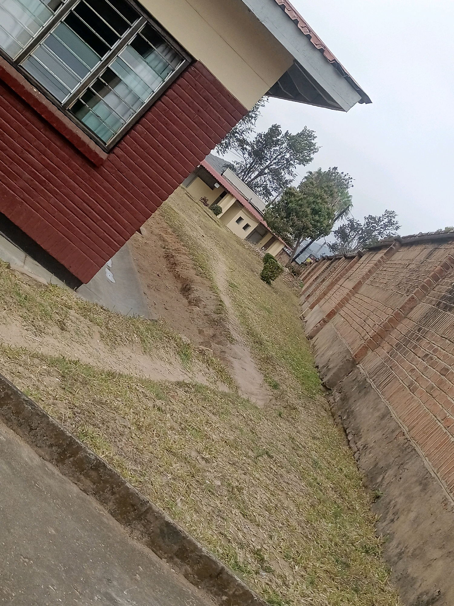This is an angled, outdoor, eye-level medium shot of a building in Limbe, Malawi. The building appears to be a school or institutional structure, with a red brick lower level and a light-colored upper level. The building has a row of windows with metal grates on the lower level. A portion of the roofline is visible, with what appears to be cracked tiles. Adjacent to the building is a steep, grassy slope leading down to a concrete ditch. On the right side of the image, a brick wall with a textured, patterned surface runs parallel to the ditch. The sky is overcast and a light gray. Trees are visible in the background, behind the building. There are no people visible in the image.
