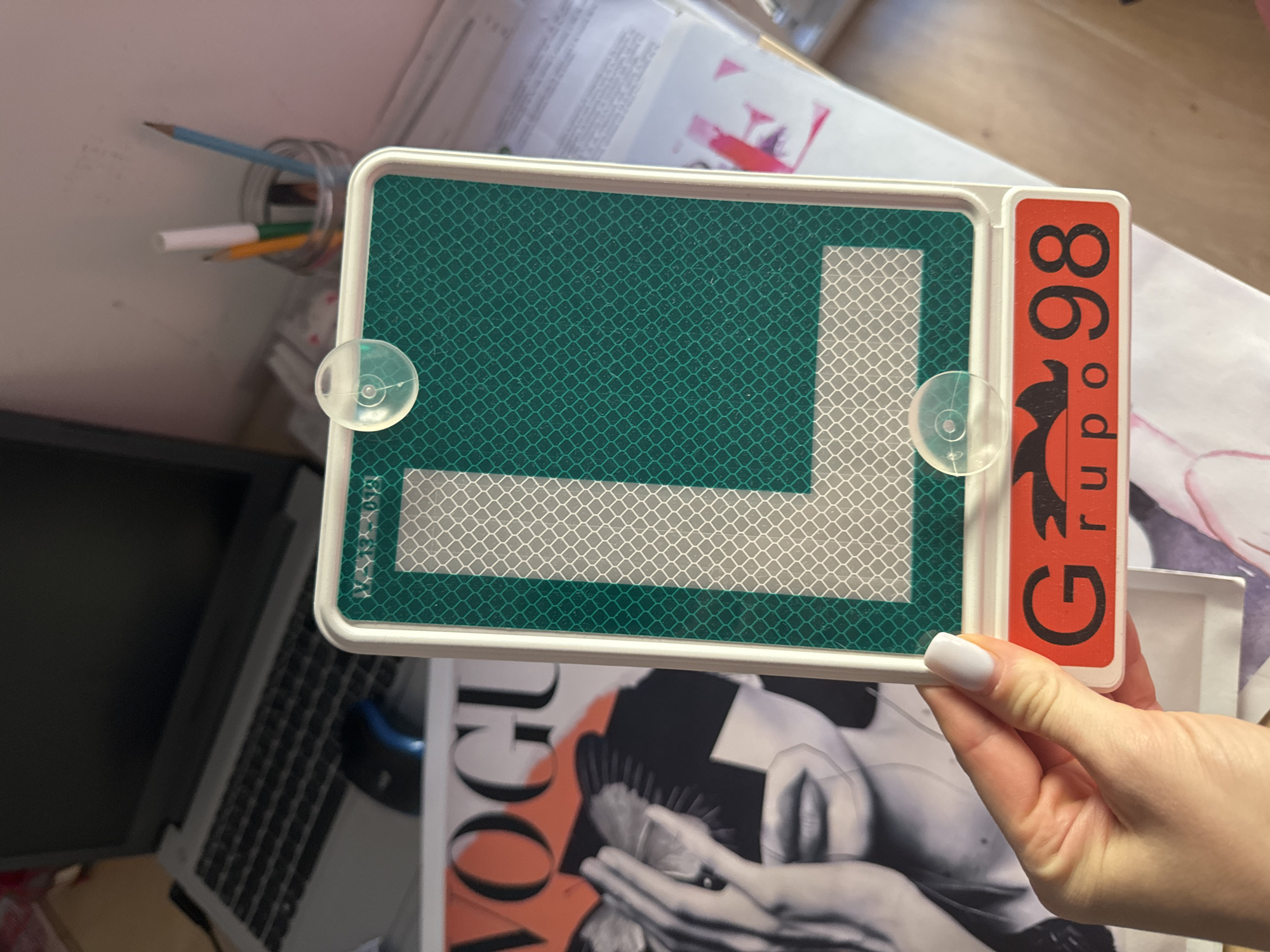 A person's hand with white nail polish holds a green and white reflective "L" sign with a white frame. The sign has two suction cups attached to it. Below the "L" sign is a red rectangle with black text that reads "Grupo 98". In the background, a laptop, a magazine cover with the word "VOGUE" on it, and a jar of pens are visible. The scene appears to be indoors, on a desk, with a wooden floor visible at the bottom. There is no indication of the city of Murcia, Spain.