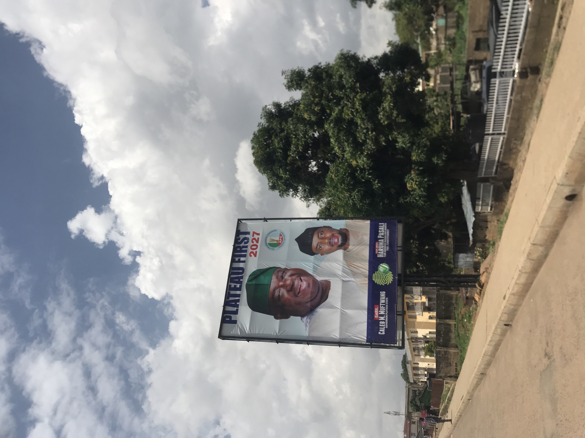 The image shows a billboard in New Stadium, Nigeria. The billboard displays "PLATEAU FIRST 2027" at the top, with images of two men. The man on the left is larger and appears to be wearing a green cap. The man on the right is smaller and wearing a dark cap. Below the images, there is text indicating "BARR. CALEB M. MUFTWANG GOVERNOR PLATEAU STATE" and "HON. BASIL M. HARUNA FOR MEMBER JARAHAU CONSTITUENCY APC". The billboard is supported by a metal frame and is situated outdoors, with a cloudy sky in the background. To the right of the billboard is a large, leafy tree. In the foreground, there is a paved road with a curb and a sidewalk. In the background, there are buildings, a fence, and some vehicles. A person is standing on the sidewalk to the left of the billboard.