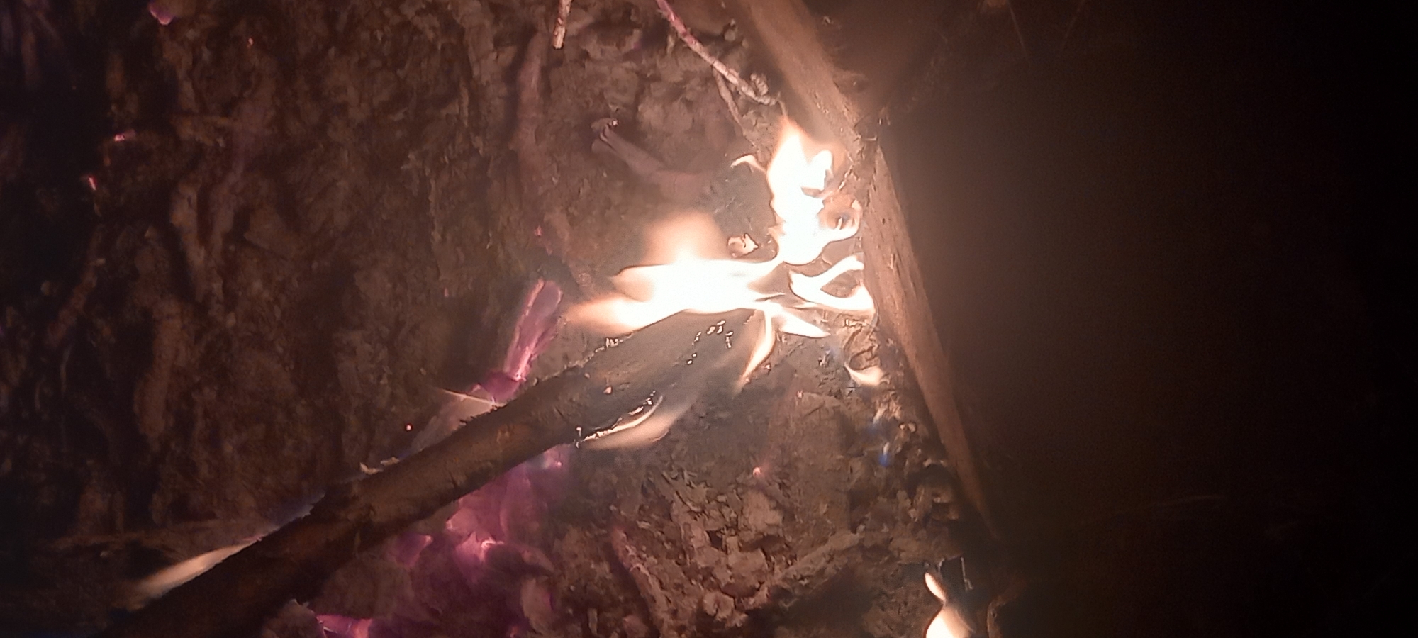 In Kryvyi Rih, Ukraine, a nighttime scene depicts a ground-level view of an active fire. The primary subject is a burning mass of wood, producing bright yellow and orange flames, with visible blue sections at the base, indicative of intense heat. Several pieces of wood serve as fuel, including a prominent elongated branch or log extending diagonally from the upper left towards the middle-right. Surrounding the active flames are glowing red and purple embers, intermingled with ash and charred debris. The fire rests on an uneven, dark ground composed of soil or earth, partially covered by ash. The scene is illuminated solely by the fire, casting light on the immediate area while the background remains in deep shadow. No individuals or specific actions are discernible.