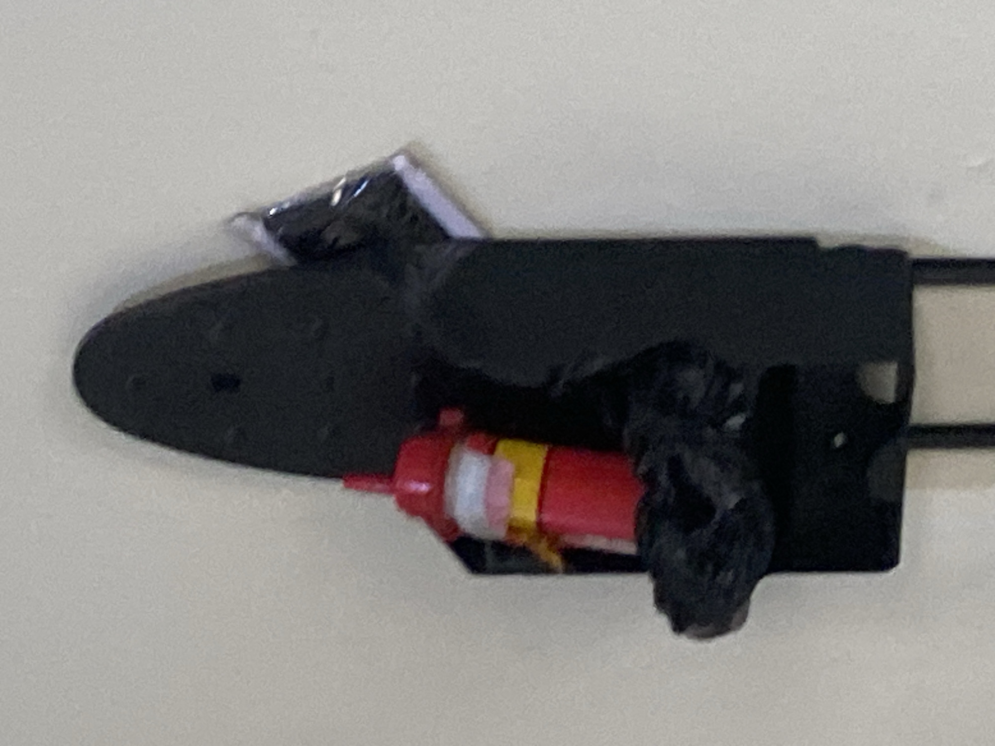 A red and yellow bottle-shaped object is secured in a black holder attached to a vertical surface, likely a wall. A small black bag is also visible within the holder. The object appears to be a fire extinguisher. The scene is indoors and illuminated by artificial light.