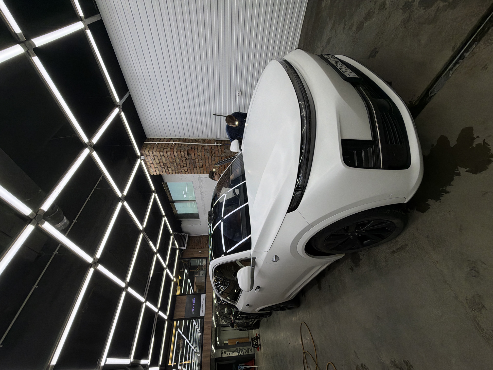 The image shows the interior of a car detailing garage. A white SUV, mostly visible from the front passenger side, is parked inside the garage. The garage has a distinctive overhead lighting grid composed of interconnected linear fluorescent lights. In the background, two individuals are visible near the car. One person appears to be examining the roof of the car, while the other stands near the car's window. The walls of the garage are gray and white. The license plate is illegible. There is a brick wall on the left side of the image. The location is Krasnoyarsk, Russia.