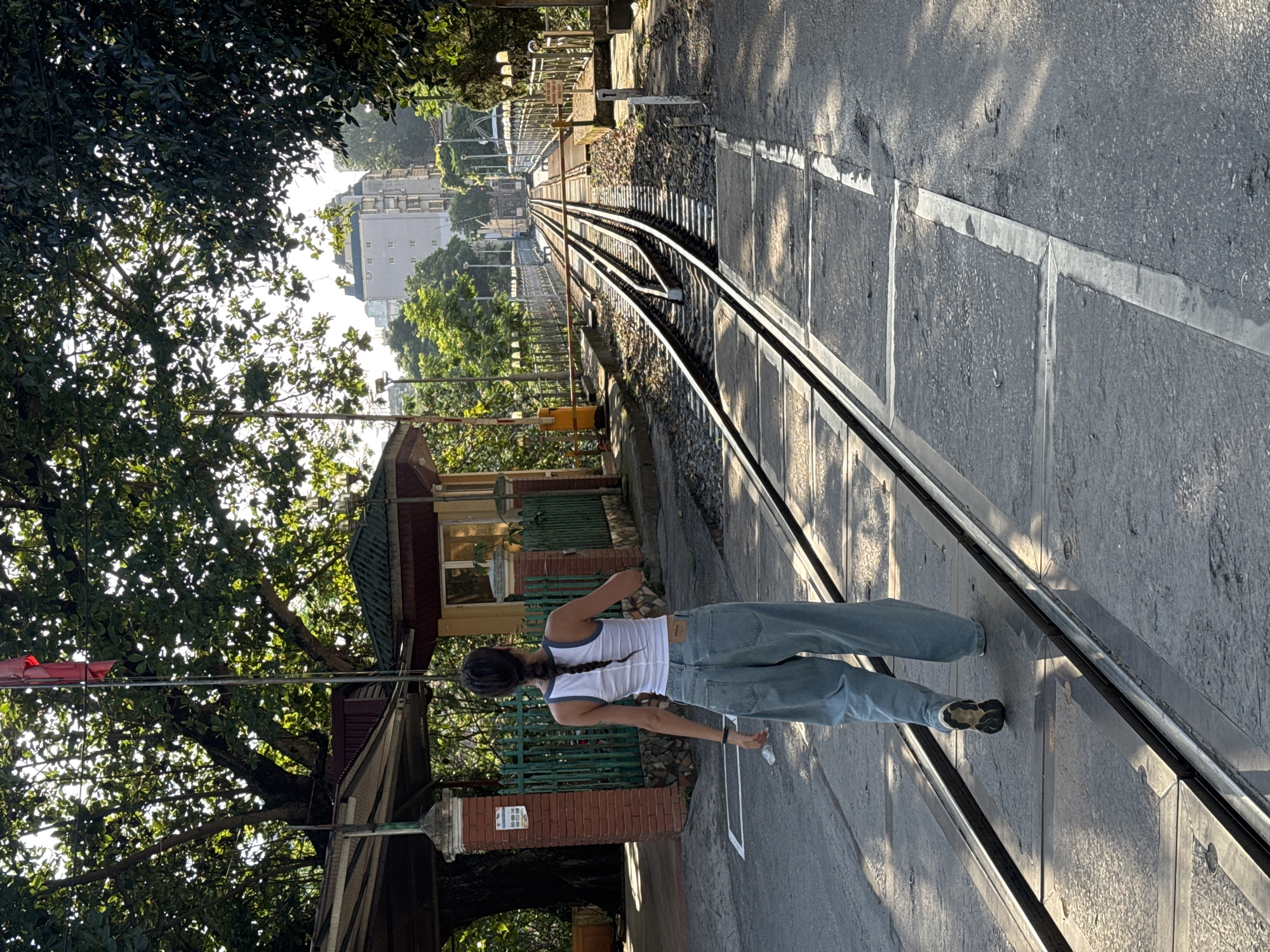 The image depicts a person standing on a narrow gauge railway track in Lao Cai Ward, Vietnam. The person, seen from the rear, is wearing a white tank top, loose blue jeans, and dark shoes. They have long dark hair styled in a braid. The person is situated on the tracks, which run parallel to a paved road. To the left, there is a verdant tree canopy and a building with a green roof. In the background, there are additional buildings and trees visible in the distance. The photograph is taken during daylight hours, as evidenced by the shadows cast on the road and railway tracks.