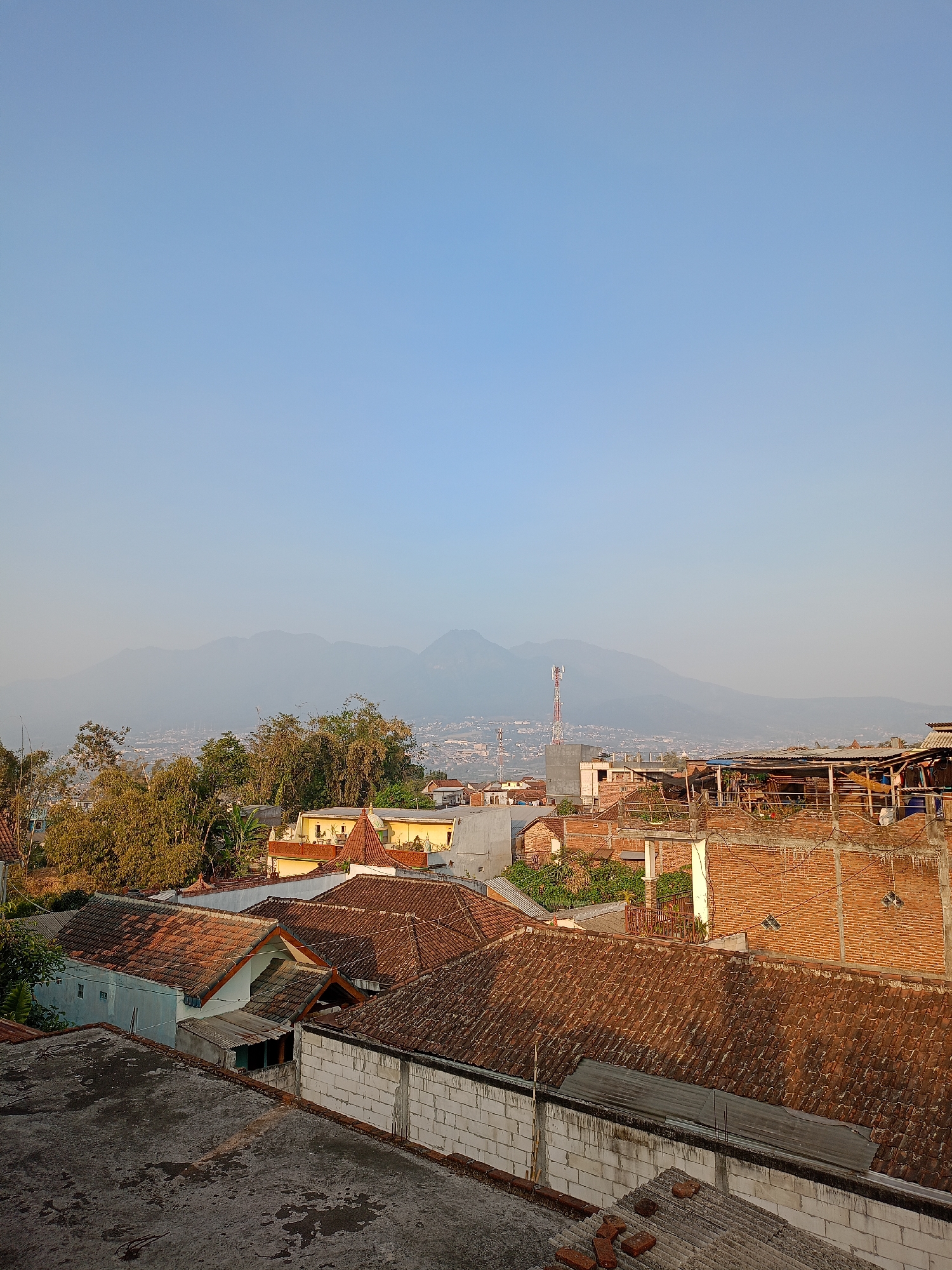 The image is an aerial, wide-angle view of a densely populated urban area, likely a residential neighborhood, set against a backdrop of hazy mountains under a clear, pale blue sky. The foreground and midground are dominated by rooftops of various structures, indicating a mix of residential and possibly commercial buildings. Many roofs are covered with traditional reddish-brown clay tiles, some appearing aged or in disrepair, while others are flat and made of concrete or corrugated metal.
The architecture suggests a tropical or subtropical climate, with exposed brickwork visible on some buildings, particularly on the right side of the image. There are also white cinder block walls and lighter colored plastered walls. Vegetation, including trees and bamboo, is interspersed among the buildings, some of which are quite dense. In the distance, a town or city is spread out, with a prominent cellular tower rising above the lower structures, and further beyond, a range of mountains is silhouetted against the sky, obscured by atmospheric haze. The lighting suggests it is either early morning or late afternoon, with the sun casting a warm glow on the rooftops. The overall impression is of a vibrant, somewhat informal urban settlement nestled in a hilly landscape. The specific location is identified as the city of Batu, Indonesia, implying a setting in East Java, known for its hilly terrain and mountainous surroundings.