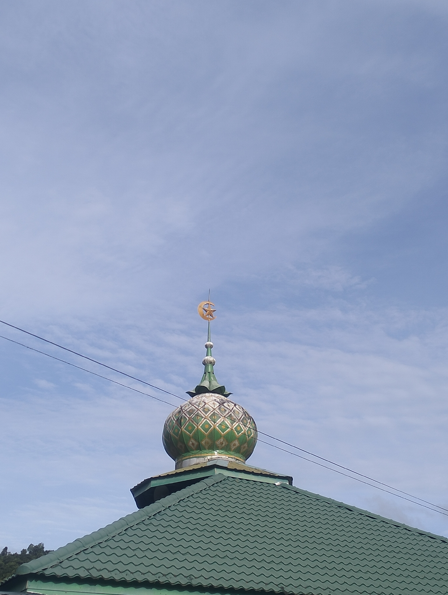 A mosque dome with a crescent moon and star finial is visible against a cloudy sky in Gleputoh, Indonesia. The dome is green and gold with a diamond pattern. The mosque has a green tiled roof. Power lines cross the sky above the mosque. Trees are visible in the background.