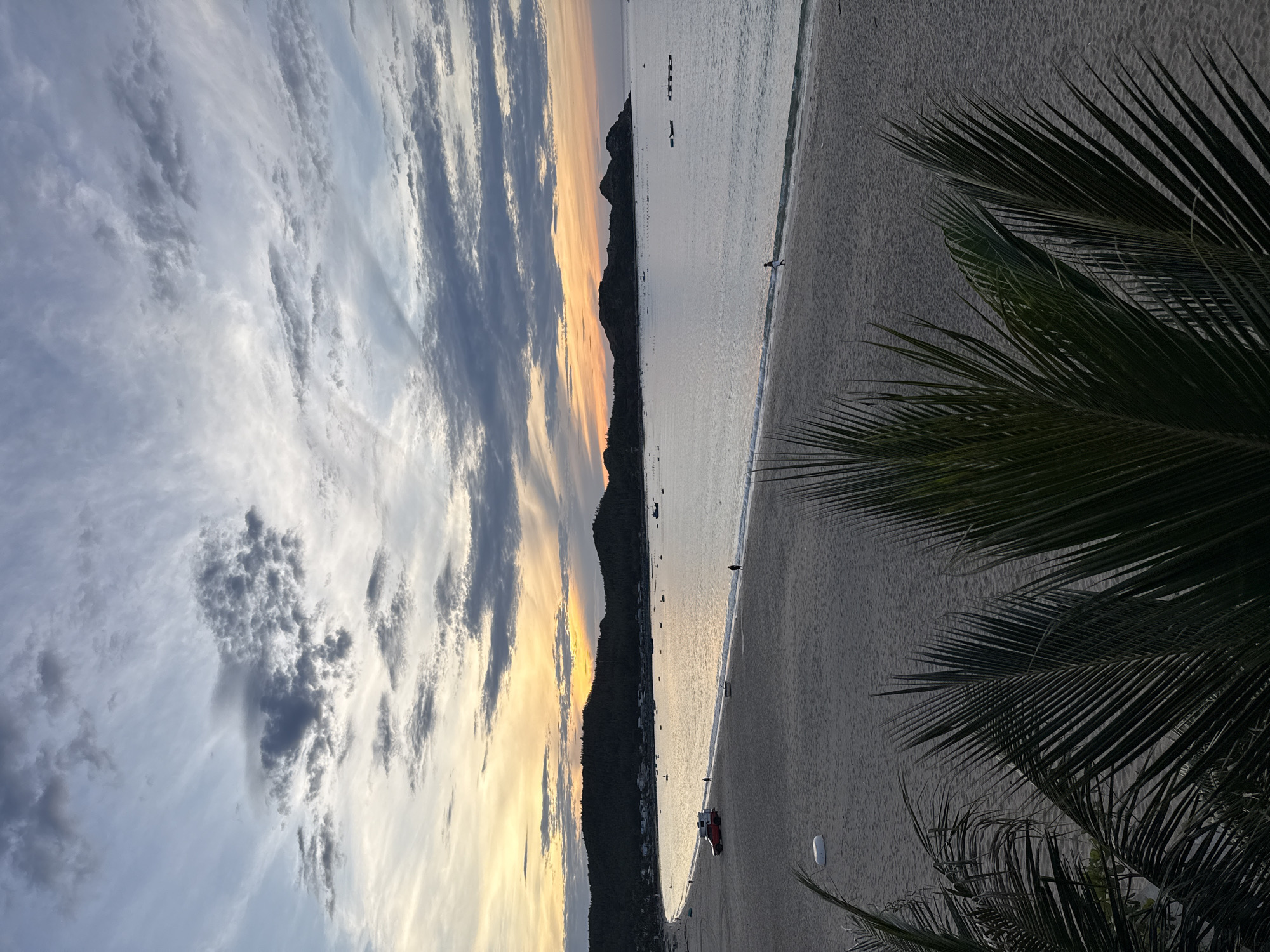 The image depicts a serene beach scene at sunset in Song Cau, Vietnam. The sky is a canvas of soft oranges, yellows, and grays, with wispy clouds stretching across the horizon. The calm sea reflects the warm hues of the setting sun, and gentle waves lap at the sandy shore. In the distance, a line of dark, tree-covered hills forms the backdrop, with a scattering of small boats and structures visible along the coastline.
On the sandy beach, a few individuals are present. One person is walking along the water's edge, while another appears to be further down the beach. A red vehicle, possibly a truck or van, is parked on the sand, and a white cylindrical object, perhaps a buoy or marker, is also visible. In the foreground, the fronds of palm trees create a natural frame, adding to the tropical ambiance. The overall atmosphere is one of tranquility and natural beauty, characteristic of the coastal areas of Song Cau.