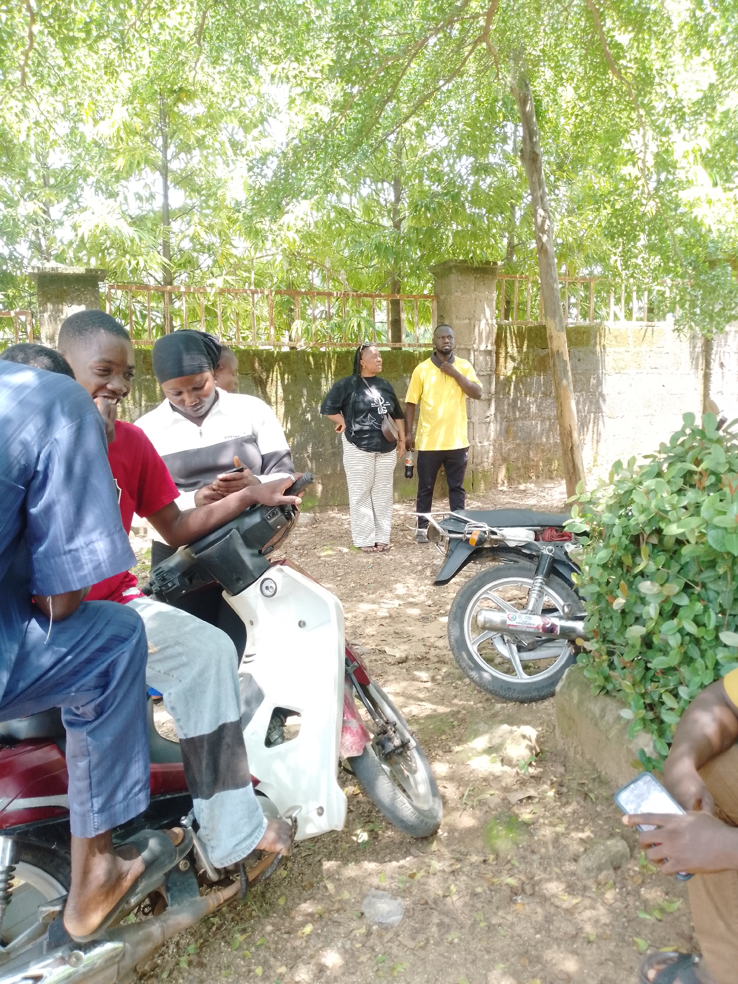 The image depicts a group of people gathered outdoors in what appears to be a casual setting. In the foreground, three individuals are positioned around a white motorcycle. One person in a blue outfit is seated, looking towards the others. Another individual in a red shirt is also seated on the motorcycle and is engaged in conversation, smiling. A third person, wearing a black headscarf and a white and black jacket, is looking down at a mobile phone they are holding.
In the background, two more people are visible. A woman in a black t-shirt and striped pants stands to the right, holding a beverage bottle. Beside her, a man in a bright yellow polo shirt and black pants is standing and appears to be speaking, gesturing with his hand. The group is situated in an area with trees providing shade, suggesting it is daytime. A stone wall and a metal fence are visible behind them. The ground is covered with dirt and fallen leaves. A second motorcycle, darker in color, is parked to the right of the group.
The overall mood seems relaxed and social. The lighting indicates it is likely midday. The presence of the motorcycles and the casual attire suggest a possible informal gathering or a break during travel.