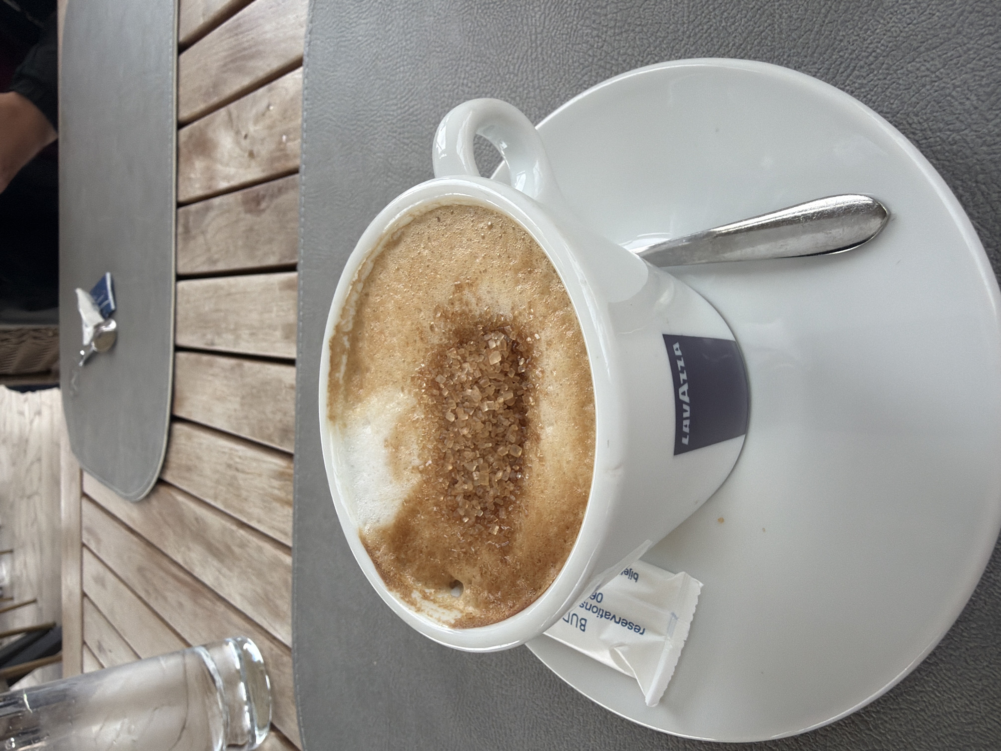 The image shows a cup of coffee on a white saucer, with a spoon resting on the saucer, taken at a table likely in Budva, Montenegro. The coffee is topped with foam and brown sugar crystals. The cup has a "Lavazza" logo on it. A white sugar packet is also visible on the saucer. The table surface consists of grey placemats and light brown wooden planks. A partial view of a glass water bottle is also present. Part of someone's arm and clothing is visible at the left side of the frame. The overall setting suggests an outdoor cafe or restaurant.