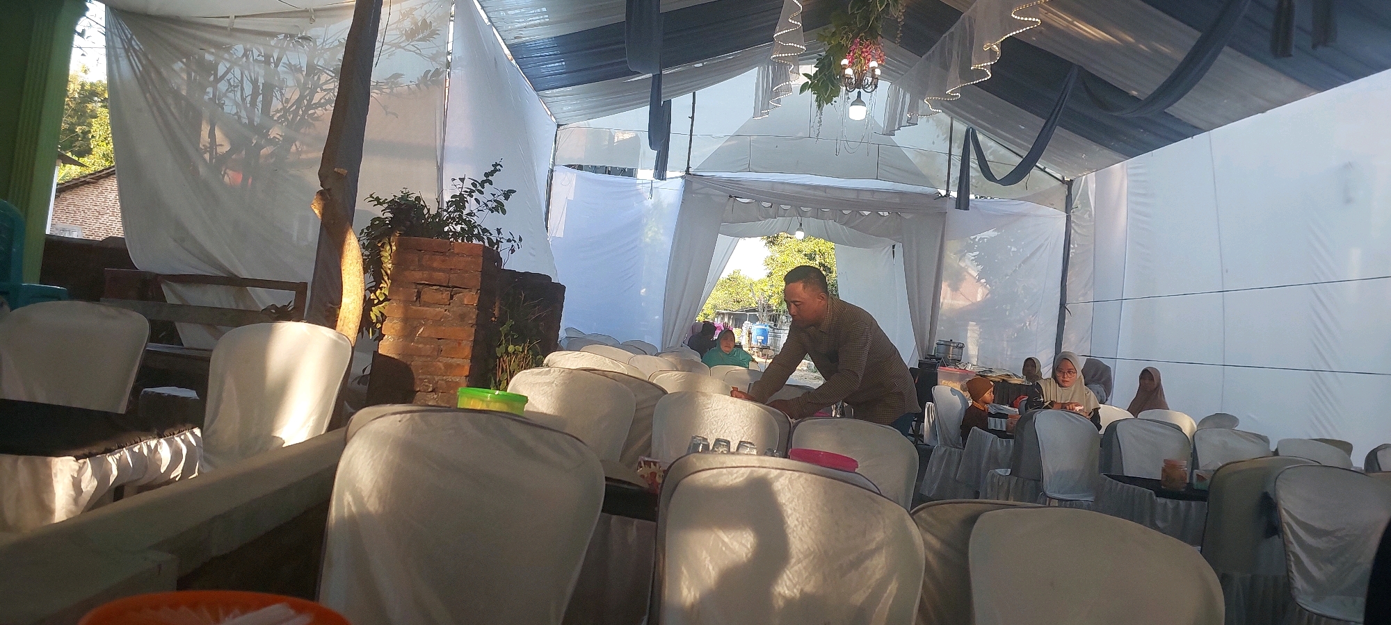 The image captures an outdoor event set up under a tent in Bukur, Indonesia. White fabric drapes the tent structure, creating an indoor-like ambiance. Several white-covered chairs are arranged in rows, some with black seat covers. A man in a patterned shirt is seen leaning over a chair, appearing to adjust it. In the background, a few women are seated at tables, some wearing hijabs. The scene suggests a gathering or a reception. Outside the tent, trees and a building with a brick wall are visible. A decorative chandelier hangs from the tent ceiling. The lighting indicates it is daytime, with sunlight casting shadows.