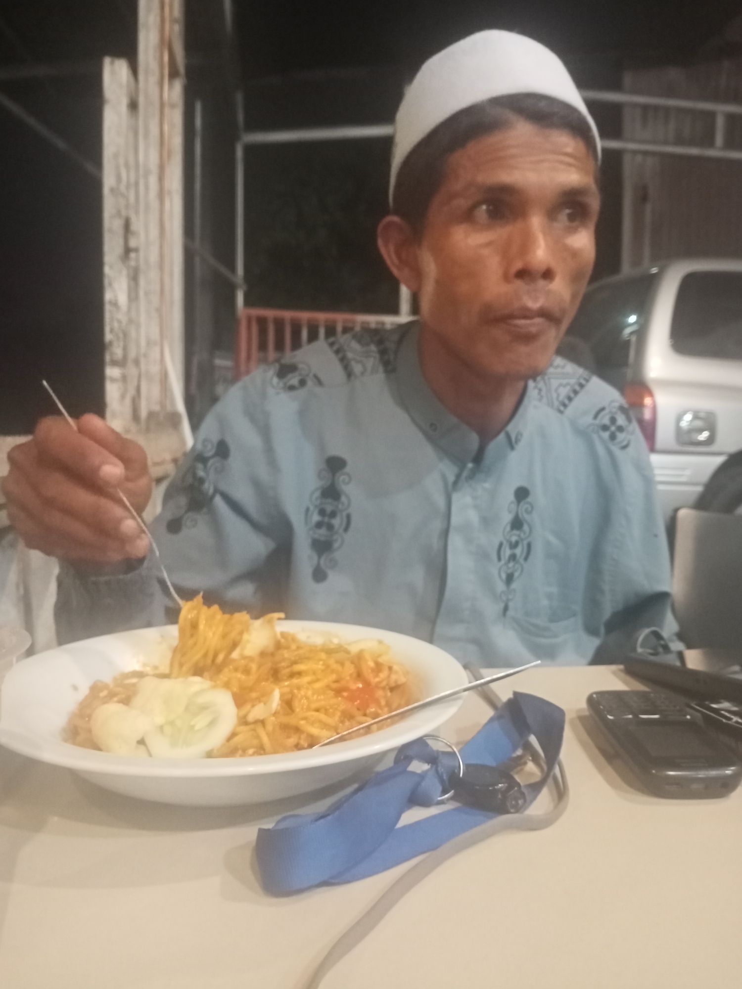 A man wearing a white cap and light blue patterned shirt is seated at a table and eating noodles with a fork. The food is served in a white bowl with what appears to be a cucumber slice on top. There is a silver spoon near the bowl. A blue lanyard with keys and a black cell phone are on the table to the right of the bowl. A silver vehicle is visible in the background. The location is identified as Tanjonghajimuda, Indonesia.
