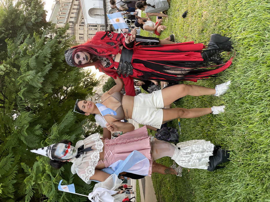 Three people in costume pose on grass in front of a building. The person on top wears a red and black outfit with dark makeup, holding an Argentina flag. The middle person wears white shorts and a bra top with an Argentina flag. The bottom person is dressed as a clown in a pink and white dress with an Argentina flag. Other people are visible in the background.