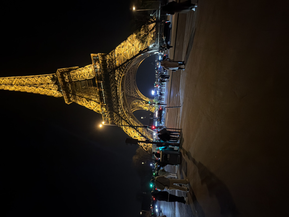 The image depicts the base of the Eiffel Tower in Paris, France, at night. The iconic wrought-iron lattice tower is prominently featured in the background, illuminated with golden light against a dark sky. In the foreground, a paved pedestrian area is visible, occupied by numerous individuals.
Approximately fifteen to twenty people are scattered across the pedestrian zone. On the left, a group of five to seven individuals stands near a dark utility box and a traffic light pole displaying red and green signals. One person in a light-colored jacket and trousers stands with their back to the viewer. To the right of this group, another person in a beige coat faces right. Further right, closer to the center, two individuals, one in a light-colored coat, stand near the edge of the pedestrian area. In the mid-ground, a street runs horizontally across the frame, populated by moving vehicles, including dark sedans and an SUV, some with active headlights. Another green traffic light is visible further down the street to the right. Beyond the street, more pedestrians are visible, along with dimly lit trees and additional street lighting. The overall scene is an urban environment, bustling with human activity and traffic under artificial illumination at night.