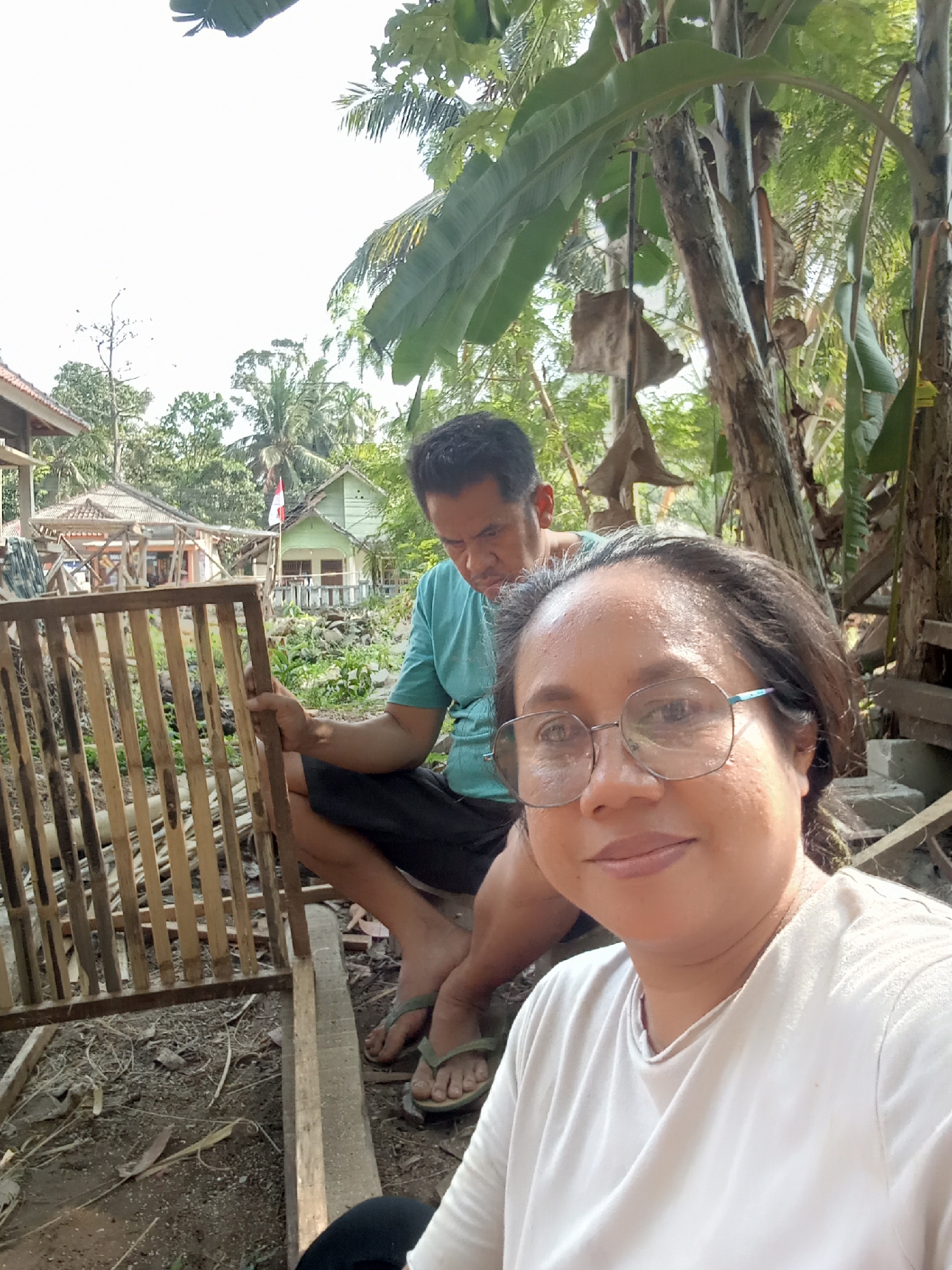 In Muara, Indonesia, a selfie captures a woman in the foreground wearing glasses and a white t-shirt. Behind her, a man in a blue t-shirt and black shorts is partially visible, holding a wooden structure. The background reveals a rural setting with houses and lush vegetation, suggesting a residential area on the outskirts of the city. The lighting indicates it is daytime.