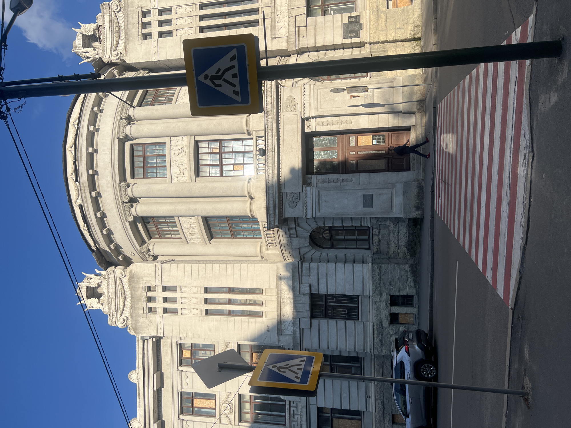 A pedestrian crossing with red and white stripes is visible in the foreground, leading towards a large, ornate building with classical architectural elements. The building appears to be made of stone and features columns, decorative carvings, and arched windows. Some windows are boarded up, suggesting ongoing construction or damage. A street sign indicating a pedestrian crossing is mounted on a pole to the left of the crossing. A silver SUV is parked on the street to the left of the pedestrian crossing. A person in dark clothing and red shoes is walking across the street on the pedestrian crossing. The scene is set in daylight, with shadows indicating the sun is at an angle. The sky is clear and blue. This is the building of the Kharkiv National University of Arts named after Ivan Kotlyarevsky, located in Kharkiv, Ukraine.