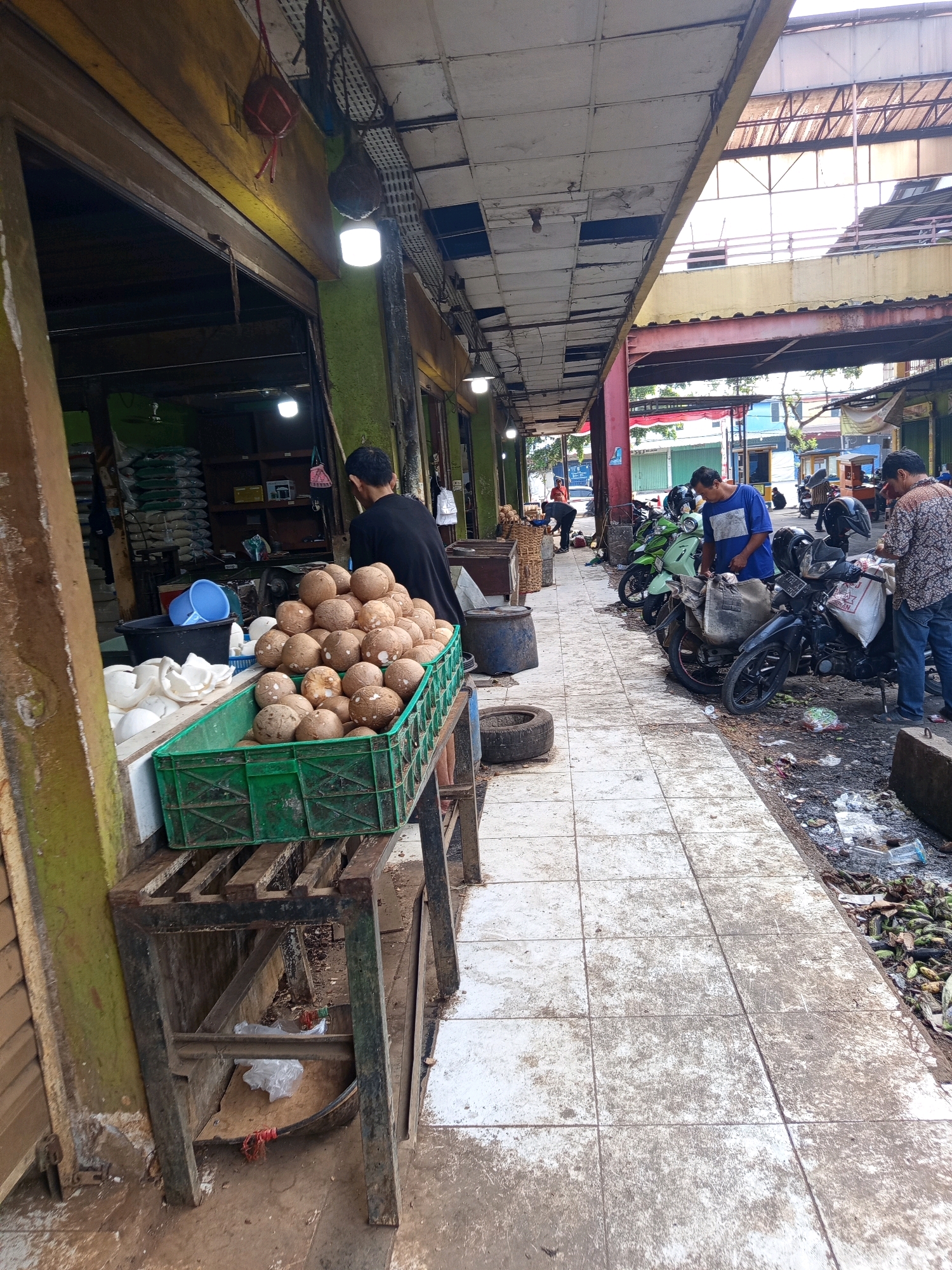 This image depicts an outdoor market scene in Karawang, Indonesia. A stall displays a large crate of coconuts, with some broken coconut shells on a table next to it. A man in a black shirt stands behind the coconuts, facing away from the camera. Further down the walkway, several motorcycles are parked. Two men are interacting near the motorcycles; one in a blue shirt is looking at a motorcycle, and another in a patterned shirt is standing next to him. The walkway is paved with tiles and appears dirty, with debris and trash visible along the edge. Buildings with awnings and open fronts line the market area, suggesting a bustling commercial environment. The overall impression is one of a typical street market, characterized by its produce display, parked vehicles, and the presence of vendors and shoppers.