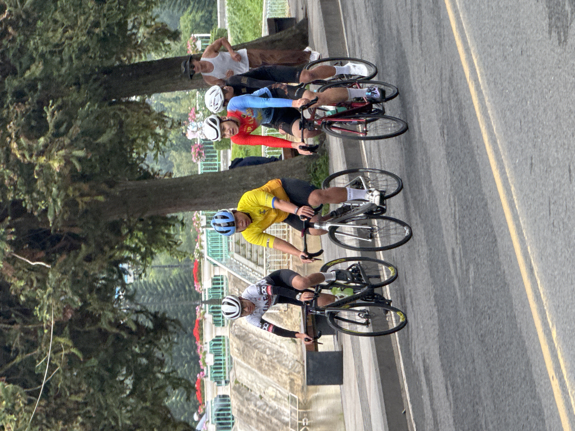 The image captures a group of cyclists participating in a race in Da Lat, Vietnam. The cyclists, dressed in athletic wear and helmets, are riding on a paved road lined with trees and decorative flower beds. In the background, a stone embankment and a body of water suggest the cyclists are near Xuan Huong Lake, a prominent landmark in Da Lat. The race appears to be well-attended, with spectators visible in the background. The overall atmosphere is one of athleticism and community engagement.