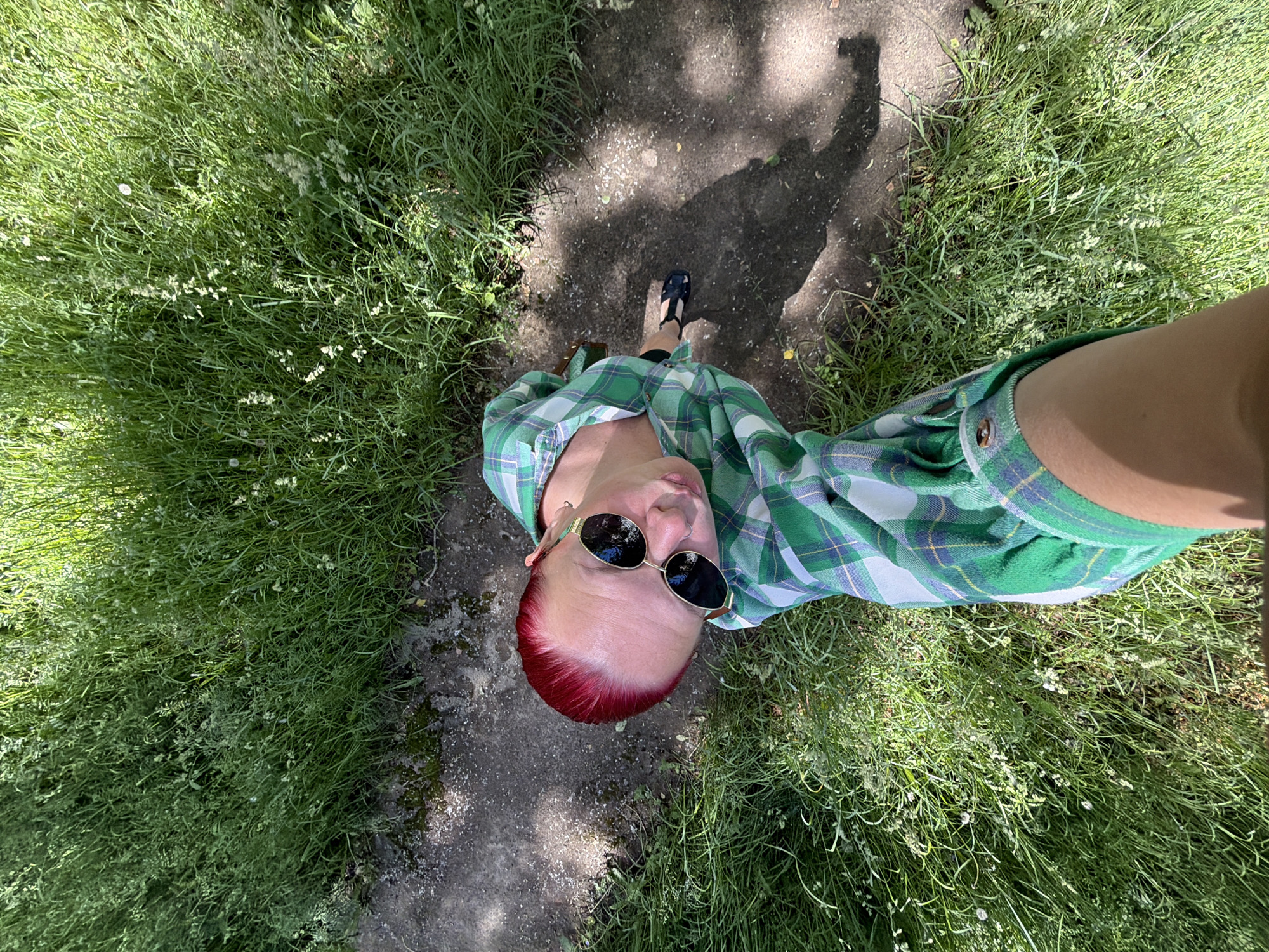 A high-angle, downward-looking image captures a single individual standing on a dirt path bordered by tall green grass in the city of Chaikovsky, Russia, under bright sunlight. The person has short, vibrant red hair and wears dark, reflective sunglasses with a gold-toned frame. They are dressed in a long-sleeved green plaid shirt with white and blue accents; one buttoned cuff is visible. A dark sandal is visible on the person's right foot, resting on the path. The narrow path is composed of packed dirt and small gravel, showing areas of direct sun exposure and elongated shadows, likely from the photographer. Dense, wild green grass, containing various plant types and some small white flowers or seed heads, extensively flanks both sides of the path.