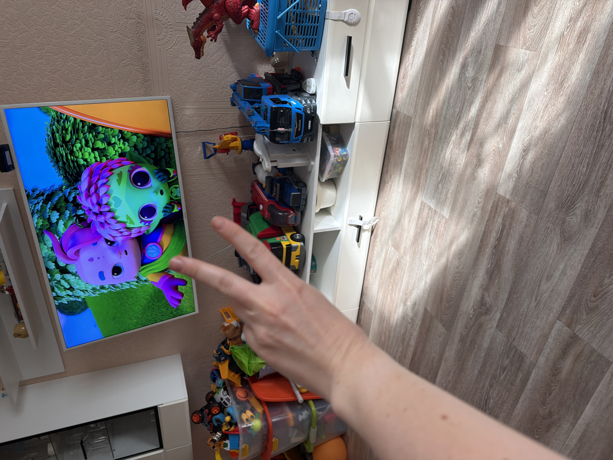 The image shows a living room with a TV screen displaying an animated cartoon. The TV is mounted on a wall above a white entertainment unit filled with various toys, including toy trucks, plastic containers, and a red dragon figure. A person's hand is visible in the foreground, holding up two fingers in a peace sign. The room has light-colored laminate flooring and the lighting suggests it is daytime, with sunlight casting shadows on the floor. The scene does not provide any specific indicators of the city of Khabarovsk, Russia.