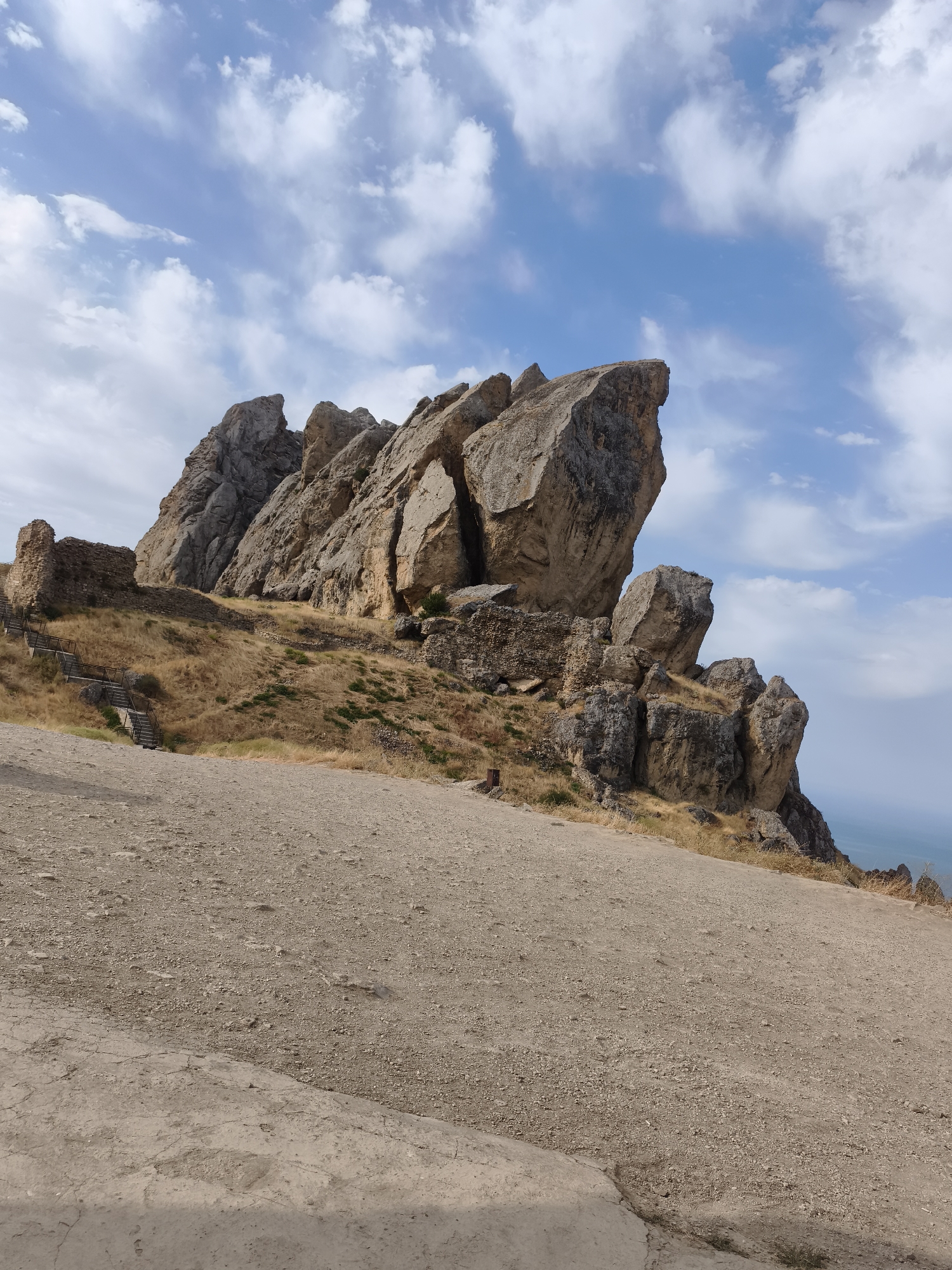 This image captures a scenic view of the ancient fortress ruins located in the village of Qalaşıxı, Azerbaijan. The fortress is perched atop a rocky hill, with a commanding view of the surrounding landscape and the distant sea. In the foreground, a dirt path leads up to the fortress, with some stone steps visible on the left side of the image. The fortress itself is a collection of crumbling stone walls and weathered rock formations, testament to its long and storied history. The sky above is a clear blue, dotted with fluffy white clouds, creating a beautiful contrast with the earthy tones of the fortress and the landscape. The dry grass covering the hill adds to the rustic charm of the scene. The overall impression is one of timeless beauty and historical significance.