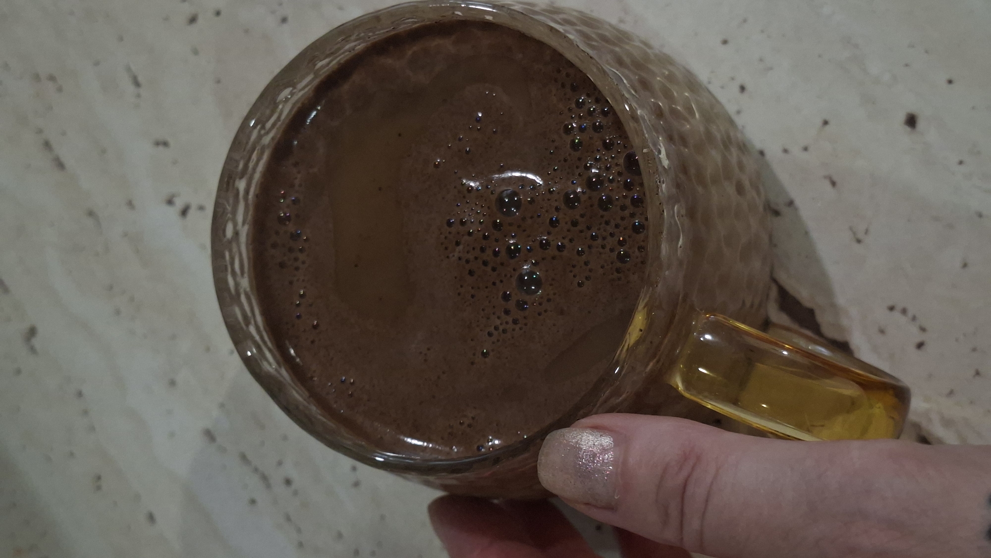 A close-up shot shows a hand with a shimmering, light-colored nail polish holding the amber-colored glass handle of a textured, clear glass mug filled with a frothy, dark brown beverage, likely hot chocolate or coffee. The mug rests on a mottled, light-colored surface that appears to be a countertop or table. The beverage's surface is covered in small bubbles, with some larger ones reflecting light. The background is out of focus, showing a textured surface. There is no indication of the city of Myadzyel, Belarus, or any other specific location within the image.