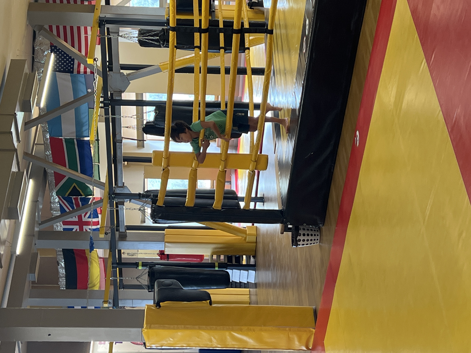 An indoor boxing ring is pictured with a young boy standing inside it. He is wearing a green t-shirt and dark shorts. The ring has yellow ropes and black padding. In the background, there are several punching bags hanging from the ceiling. Flags of different countries are displayed on the wall above the punching bags. The floor is covered with yellow and red mats. The overall scene appears to be a training facility, likely a gym or a martial arts studio. The scene is located in Almaty, Kazakhstan, specifically at the "Tiger" boxing club.