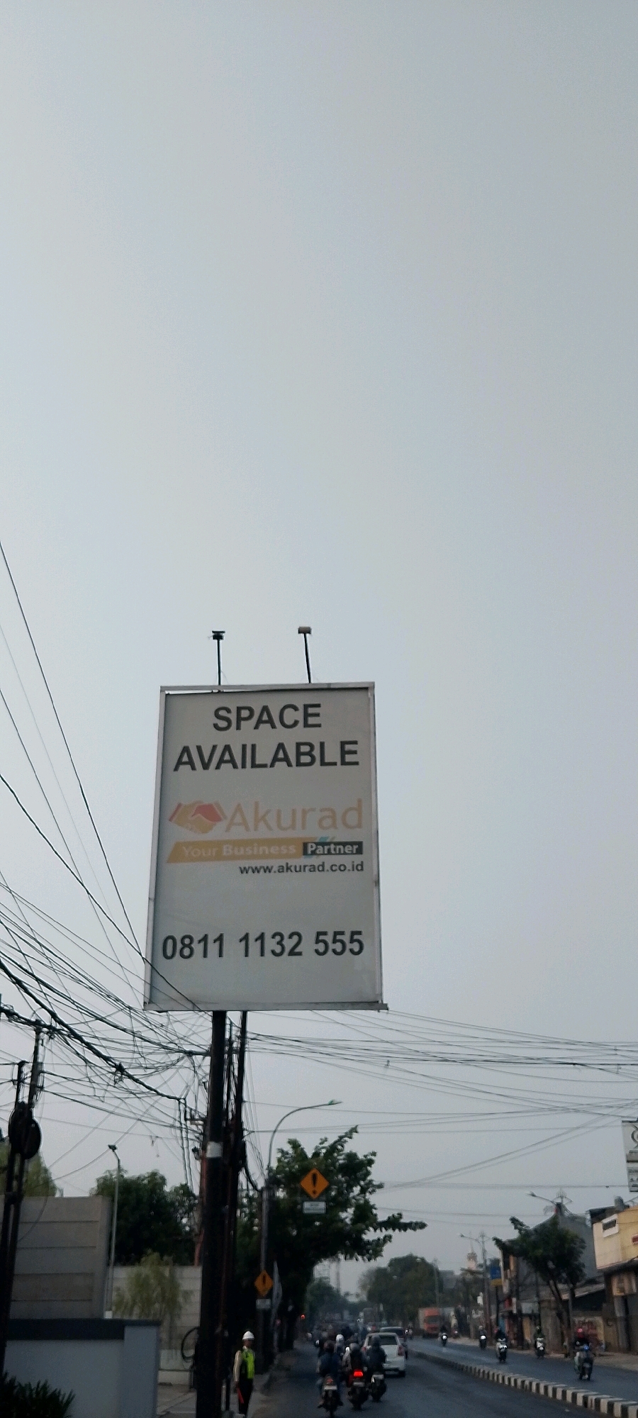A vertical, eye-level, medium shot captures a billboard advertising "SPACE AVAILABLE" for Akurad, with a contact phone number and website, positioned on a street in Bekasi, Indonesia. The billboard is mounted on a pole, partially obscured by a dense network of electrical wires overhead. Below the billboard, traffic flows along a road, with several motorcycles and cars visible. A person wearing a reflective vest and a hard hat stands near the base of the billboard pole, facing away from the camera. The sky is overcast, contributing to a muted, grey ambiance. The foreground features a grey wall on the left side of the frame, partially obscuring a building. The overall scene depicts an urban environment with commercial signage and active street life under a grey sky.