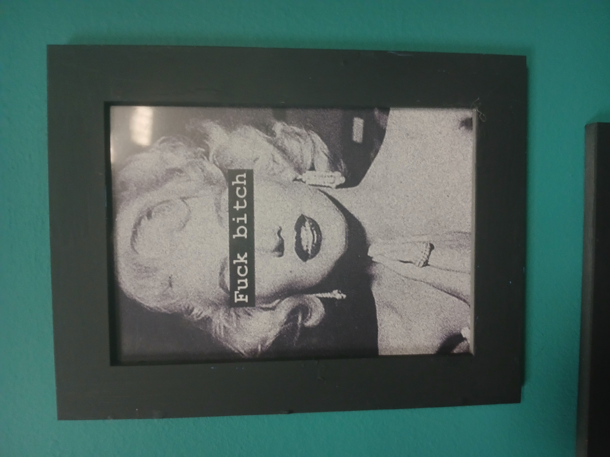 A framed black and white photograph of Marilyn Monroe hangs on a teal wall. Her face is partially obscured by a black rectangle with white text that reads "Fuck bitch". She is wearing dangling earrings and a dress with a ruffled neckline. The frame is black and appears to be made of wood. The image is taken from a slightly elevated angle.