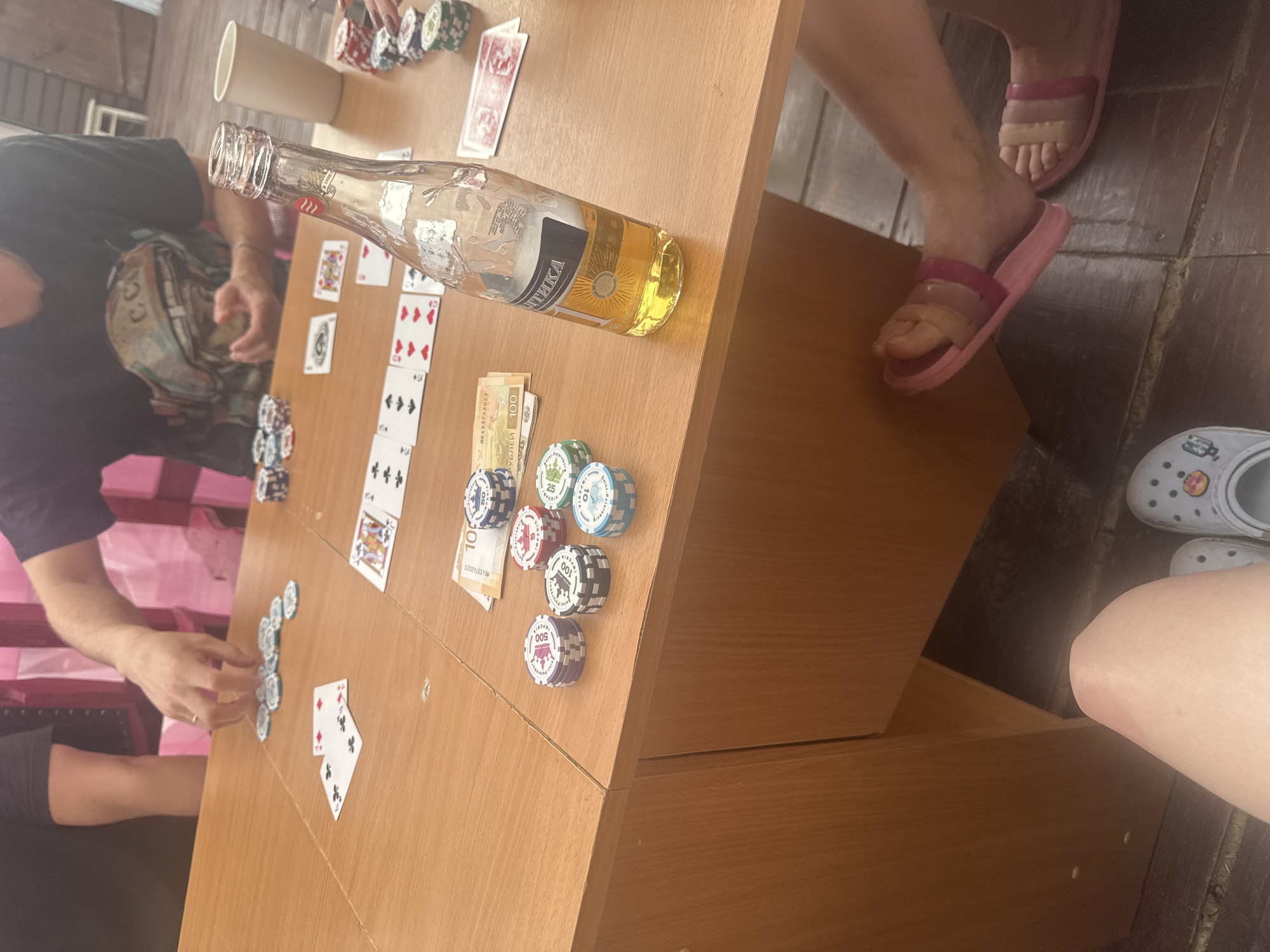The image depicts a casual gathering at a wooden table, likely a card game, with chips and cards laid out. A partially filled bottle of beer with a label reading "Балтика" is prominently placed on the table. The setting appears to be outdoors, suggested by the wooden floorboards and natural lighting. In the foreground, a person's legs are visible, wearing pink slides and white Crocs. The overall atmosphere is relaxed and informal. There is no information in the image to suggest the location is in Zhebrei, Russia.
