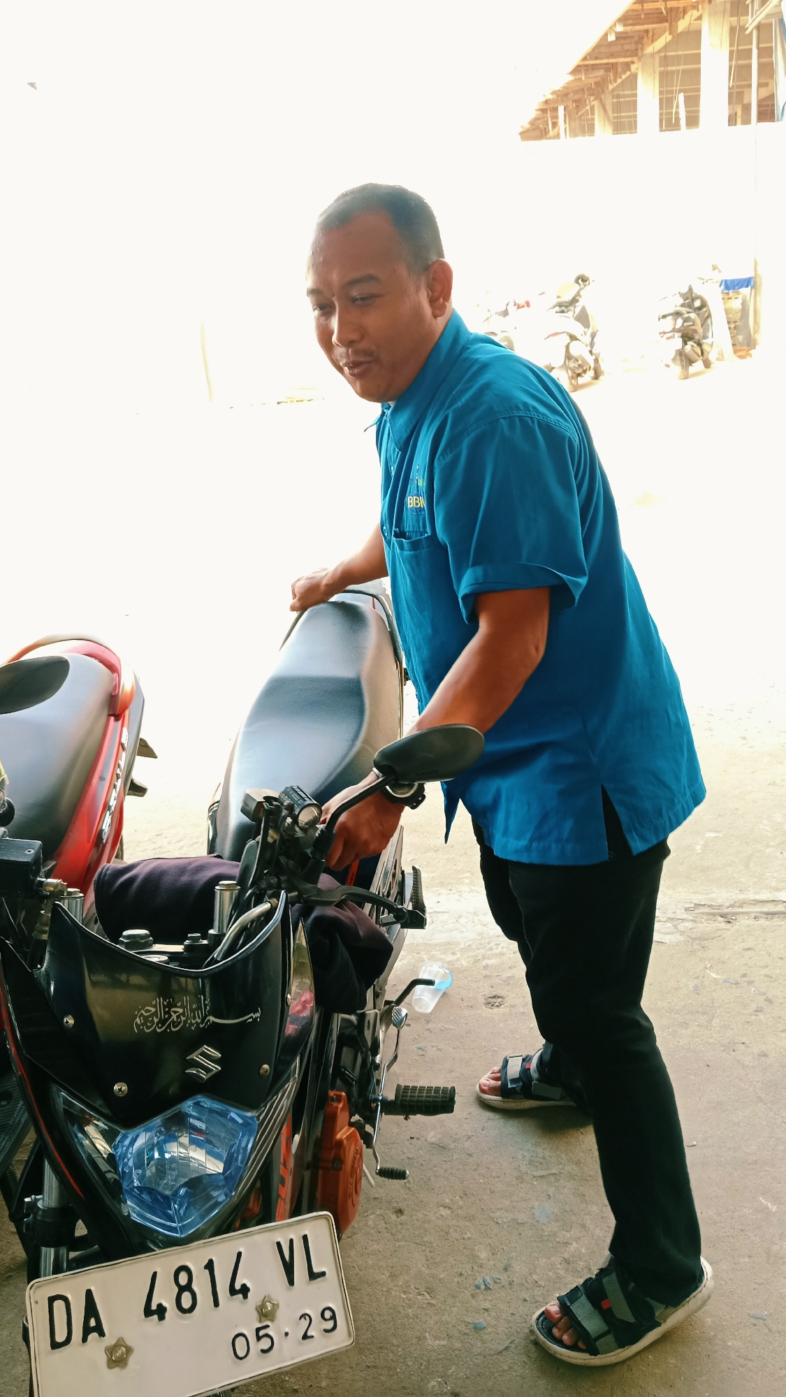 In Handil Bakti, Indonesia, a man in a blue collared shirt and black pants stands beside a motorcycle, with his right hand on the motorcycle seat. The man has short dark hair and is looking downwards, with his mouth slightly open. The motorcycle is black with red accents and has a license plate with the characters "DA 4814 VL" and "05 29". Another motorcycle, predominantly red, is partially visible to the left of the frame. The background consists of a lightly-colored wall and overhead structures, suggesting an outdoor area possibly under construction or a covered parking space. The ground is paved and appears worn. Several other motorcycles are visible in the background, some with reflective surfaces. A plastic bottle lies on the ground near the man's feet.