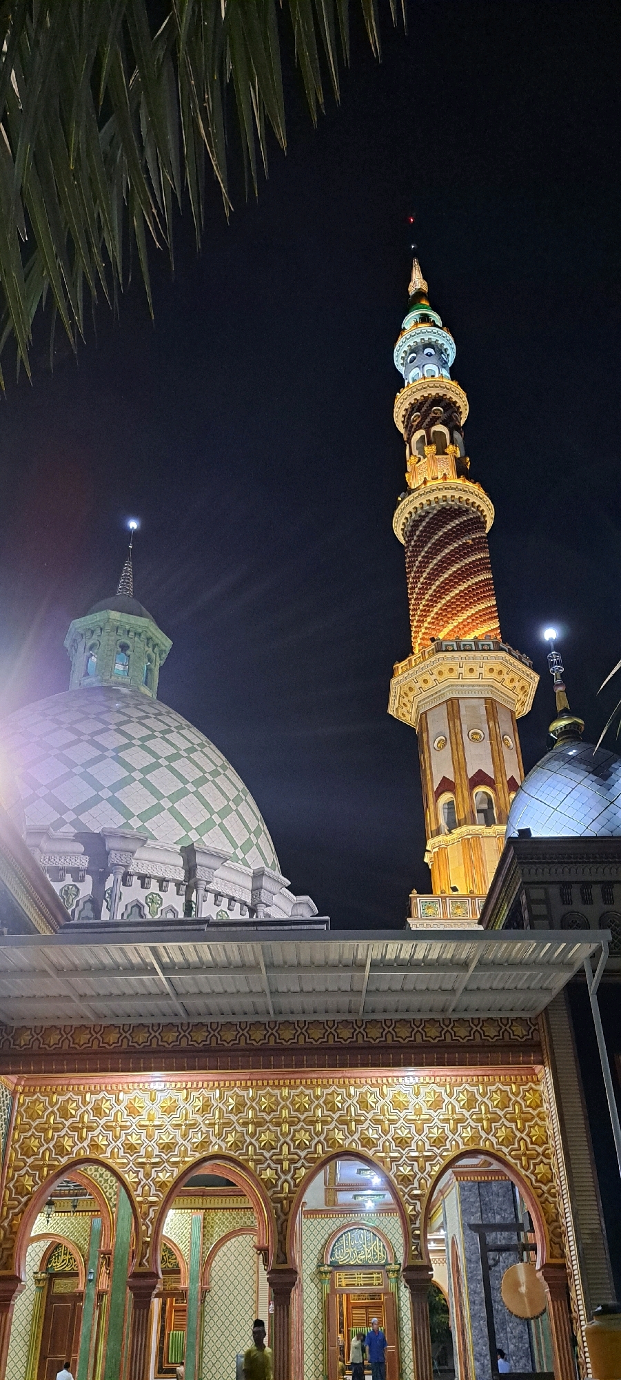 An exterior nighttime, low-angle shot shows a mosque with a large dome on the left and a tall, ornate minaret on the right. The dome is patterned with a green diamond grid. The minaret has a helical stripe design in red and gold, with decorative balconies and a pointed spire. In the foreground, there is an arched colonnade with intricate golden geometric patterns and Islamic calligraphy. A portion of a palm frond is visible in the upper left corner. Several individuals are present, two standing in the arched entryway and one in the colonnade. The scene is illuminated by artificial lights, highlighting the architectural details of the mosque. This structure is identified as the Sunan Ampel Mosque in Surabaya, Indonesia, not Baujeng.