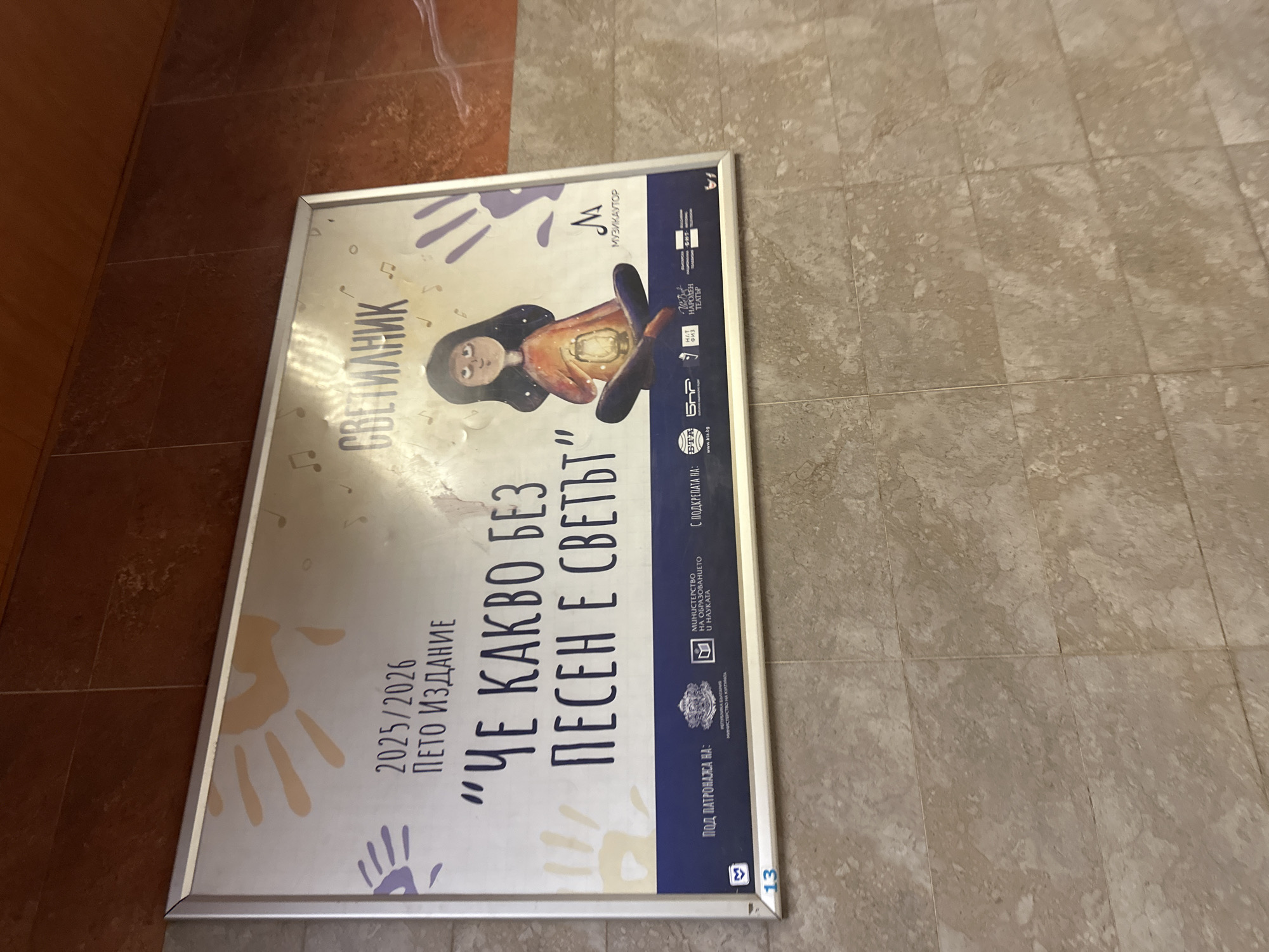 The image depicts a poster lying on a tiled floor, likely inside a building. The poster, which is contained within a silver frame, is advertising an event called "СВЕТИЛНИК" or "SVETILNIK." The text on the poster indicates it is the fifth edition (ПЕТО ИЗДАНИЕ) of this event and includes the phrase "ЧЕ КАКВО БЕЗ ПЕСЕН Е СВЕТЪТ," which translates roughly to "What is the world without a song?" The poster displays a stylized illustration of a person holding a glowing object. It has the logos of Музикаутор (Musikautor), Народний театр (National Theatre), BTA, and the Ministry of Education and Science. Based on the presence of Bulgarian text and logos, it can be deduced that the location is likely in Bulgaria, and because it is the "National Theater," it is most likely the capital city, Sofia.