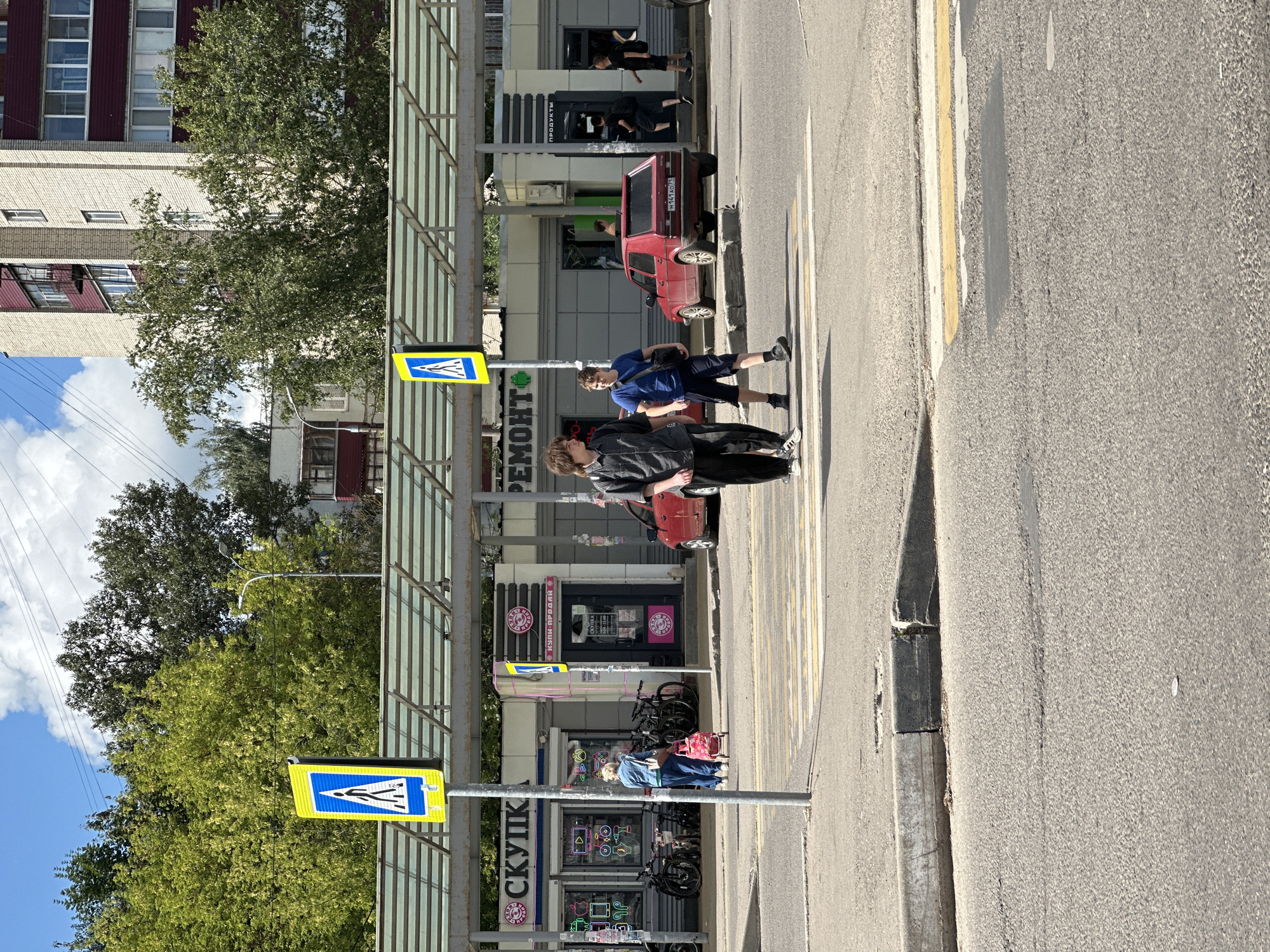 The image captures a street scene in Chekhov, Russia, on a sunny day with scattered clouds. In the foreground, a pedestrian crossing is visible, marked by white and yellow stripes. Two young men are crossing the street from right to left. The man on the left has dark, curly hair and wears a black shirt and black pants. The man on the right has brown hair and wears a blue t-shirt and dark shorts, carrying a black bag over his shoulder.
Behind the pedestrian crossing, a row of shops is situated under a covered walkway with a translucent roof supported by grey beams. A pedestrian crossing sign, a blue square with a white silhouette of a person walking, is mounted on a pole to the left of the walkway. Another similar sign is visible further down the walkway. One of the shops has the word "СКУПКА" (SKUPKA) displayed prominently in pink lettering, and another has "РЕМОНТ" (REMONT) and "ПРОДУКТЫ" (PRODUCTS) visible. Bicycles are parked near one of the shops.
Two red cars are parked on the right side of the street. The car closer to the pedestrian crossing is a hatchback. In the background, a multi-story residential building with light-colored brick and red accents rises against the sky. Trees with green foliage are scattered throughout the scene, some lining the street and others behind the buildings. A lamppost is also visible. The overall environment suggests an urban setting in Russia, likely a commercial or mixed-use area.
