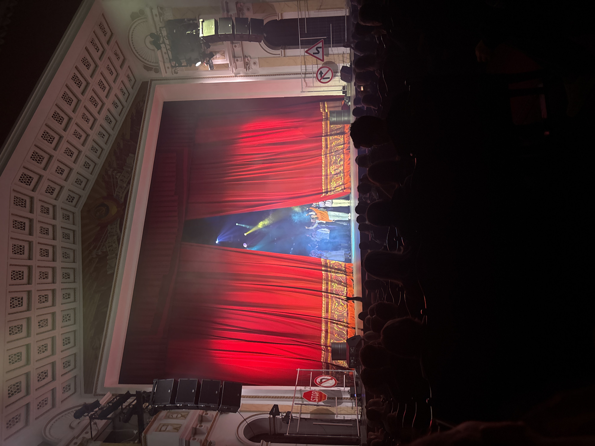 A partial view of an audience is seen in dim light, observing a stage performance with a red curtain opening to reveal performers lit by stage lights. Above the stage are speakers and stage rigging. Road signs are placed on the stage near the curtain edge. The architectural details of the theatre's walls and ceiling are partially visible. This is likely a scene from the Perm Opera and Ballet Theatre in Perm, Russia.