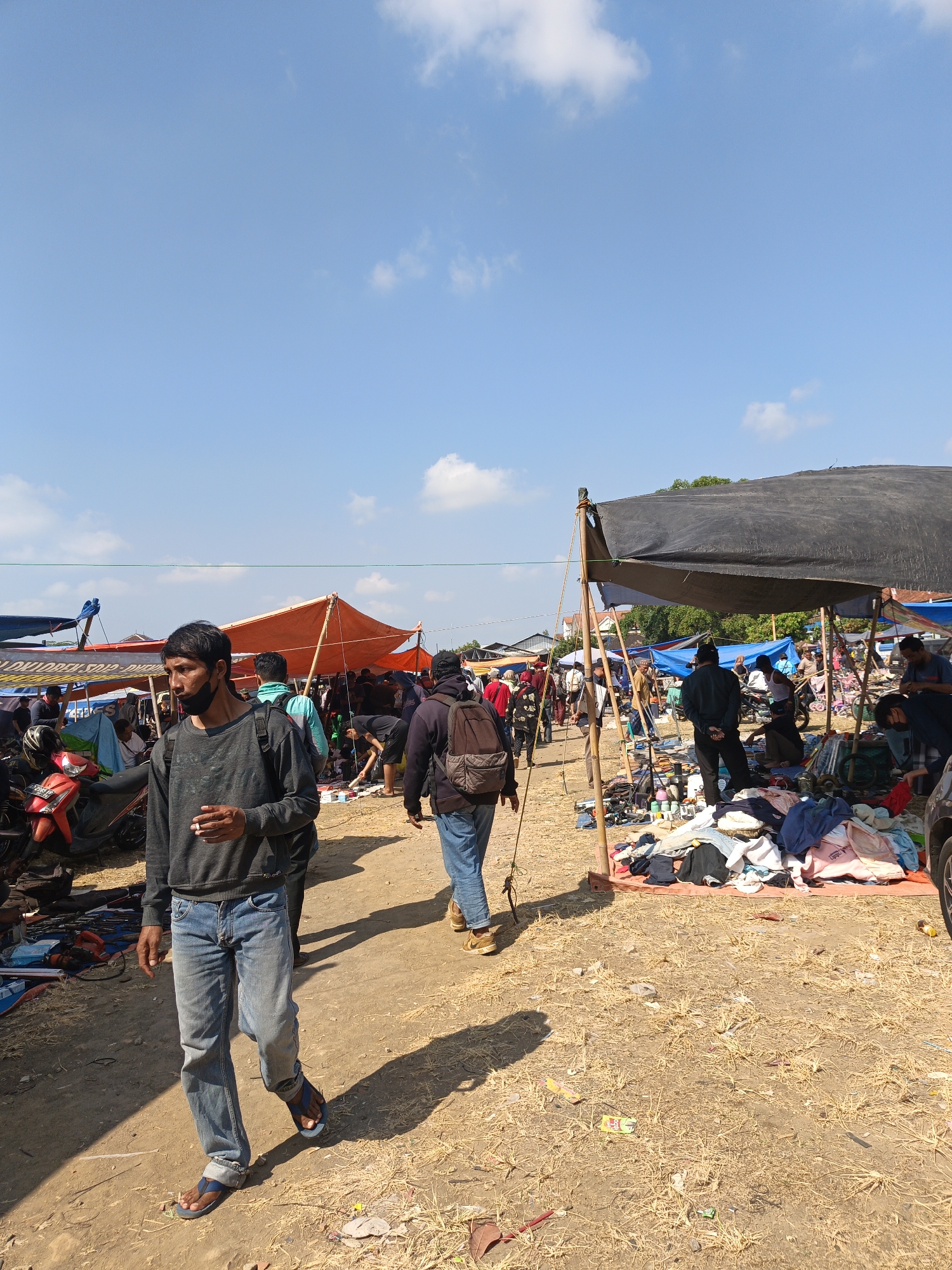 This is a daytime, eye-level, full shot of a busy outdoor market in Jatinom, Indonesia. The sky is a clear, bright blue with scattered white clouds. In the foreground, a man wearing a dark gray long-sleeved shirt and blue jeans walks away from the camera, holding a cigarette in his right hand. He is wearing blue sandals. Behind him, other people are visible browsing through stalls set up under tents.
The market appears to be a flea market or a secondhand goods market. Tents are erected to provide shade, some with orange tarps and others with black tarps. Various goods, including clothing, are displayed on the ground and on tables. Motorcycles are parked to the left of the frame. The ground is covered with dry grass and dirt. In the background, more tents and stalls extend into the distance, suggesting a large market area. The scene is bustling with activity, indicating a lively marketplace.