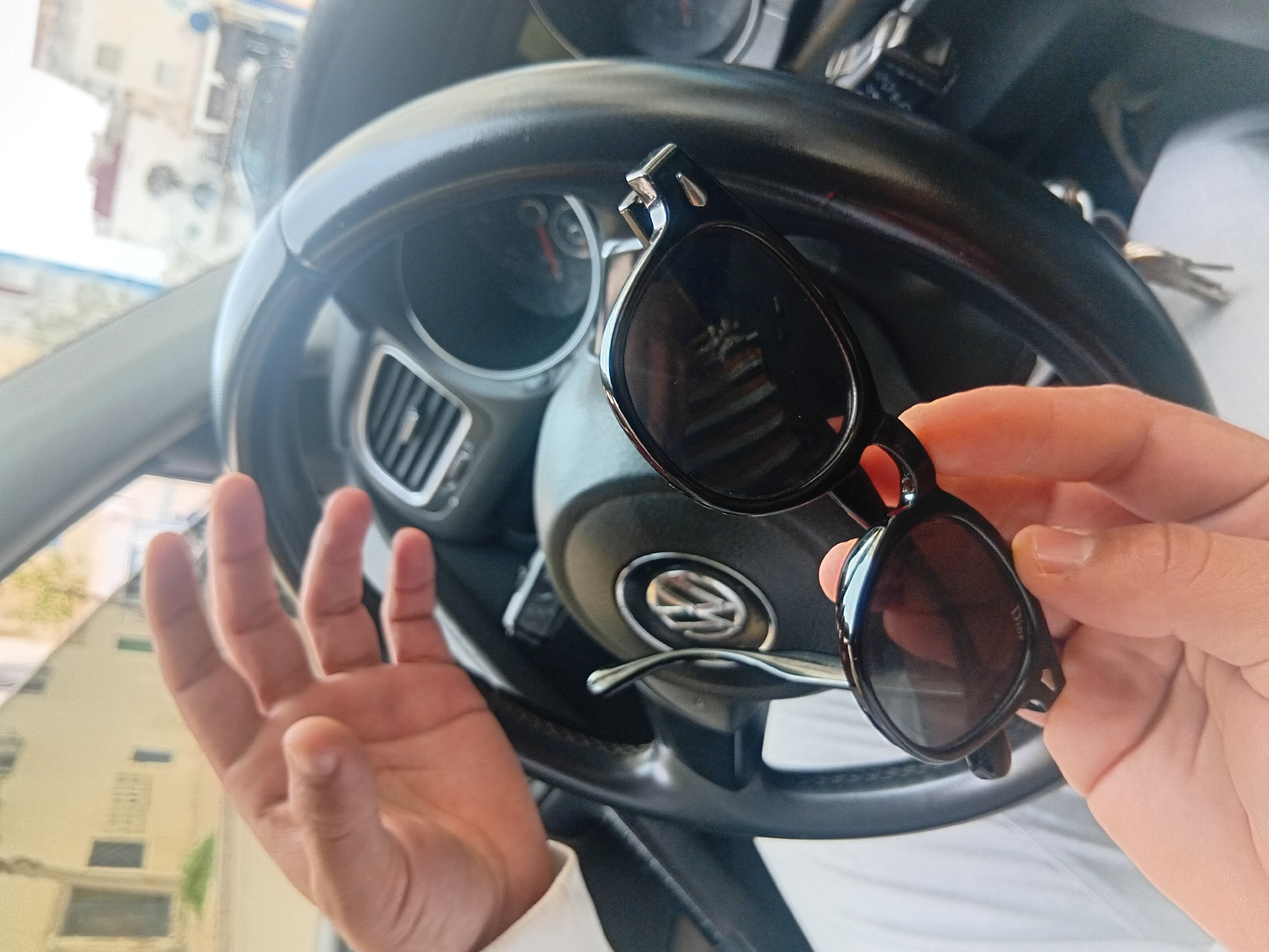 The image captures a close-up view inside a car in Martil, Morocco, during the daytime. The primary subjects are a person's hands and a pair of black-framed sunglasses. The right hand holds the sunglasses, revealing the brand "DIOR" faintly in reverse on one of the dark lenses. The left hand is open, palm up, with fingers spread.
The setting is the interior of a car, indicated by the prominent black steering wheel with a silver Volkswagen (VW) logo in the center. Parts of the dashboard, including an air vent and instrument gauges, are visible behind the steering wheel. Car keys are seen hanging near the steering column. Through the car window on the left, a light-colored building with visible windows and some distant greenery suggests an urban or suburban environment. The bright, natural light indicates a clear, sunny day. The perspective appears to be from a first-person point of view within the vehicle.
