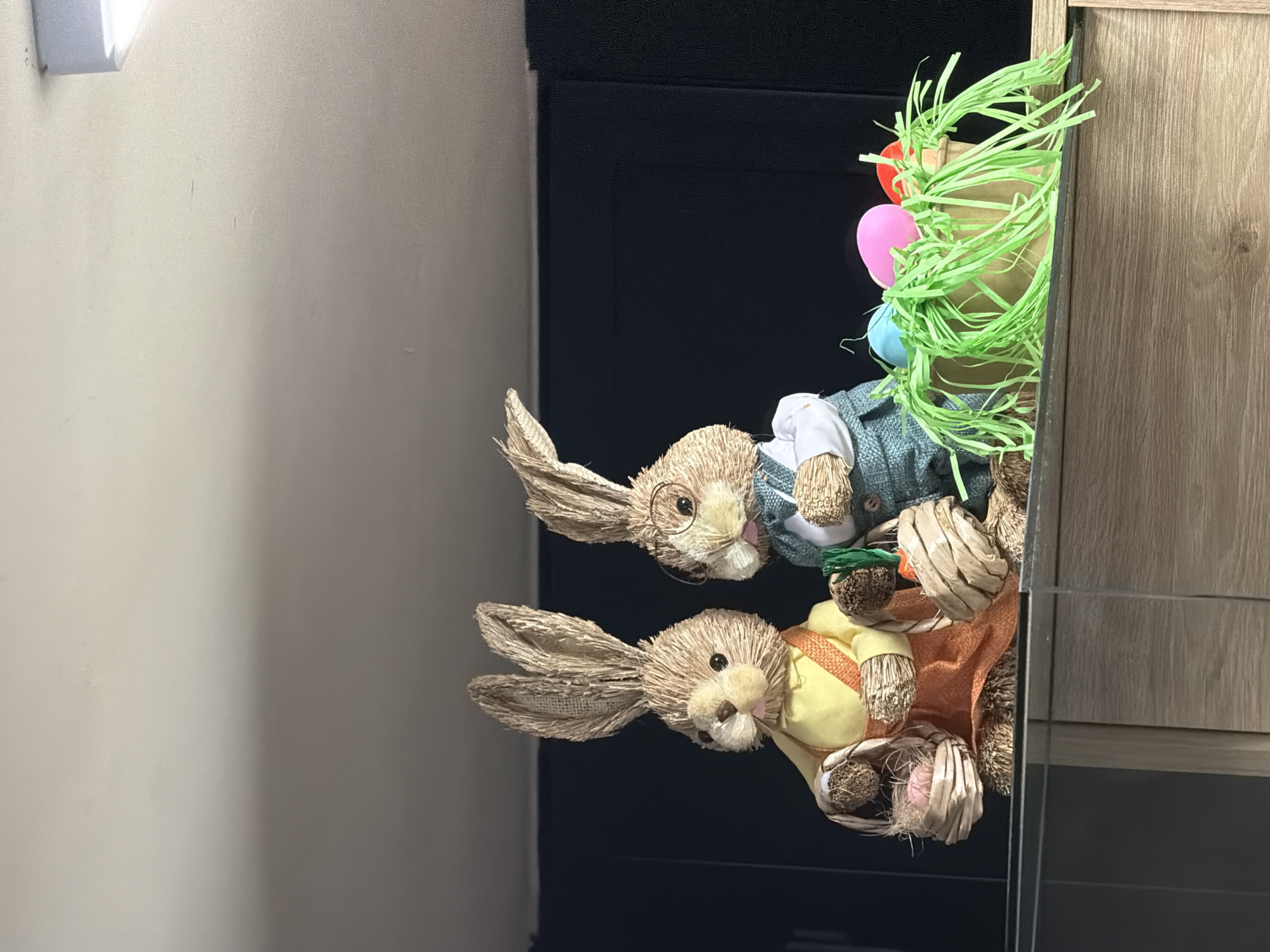The image shows two stuffed bunnies partially obscured by a vase with green Easter grass and decorated with Easter eggs. The bunnies are leaning against a dark cabinet and a wooden cabinet. The scene appears to be indoors, possibly in a home. There is no information in the image to suggest this is in Sofia, Bulgaria.