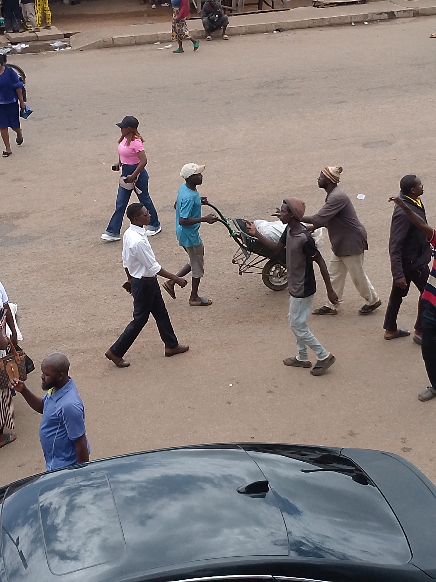 The image depicts a street scene in Bukuru, Nigeria. In the foreground, the shiny black roof of a car reflects the sky. Several people are walking on the paved street. On the left, a man in a blue polo shirt is looking to his right. Further back, a man in a white shirt and black pants is walking to the left, holding a phone in his right hand. To his right, a boy in a blue t-shirt and grey shorts is pushing a wheelbarrow loaded with white material. Behind him, two men are walking to the right; one in a brown shirt and light-colored pants, and another in a dark jacket and dark pants. A woman in a pink t-shirt and blue jeans, wearing a black cap, walks to the left. Another woman in a blue dress is walking to the left in the upper portion of the image. In the background, other pedestrians are visible. The environment appears to be an urban or semi-urban setting with buildings and a road.