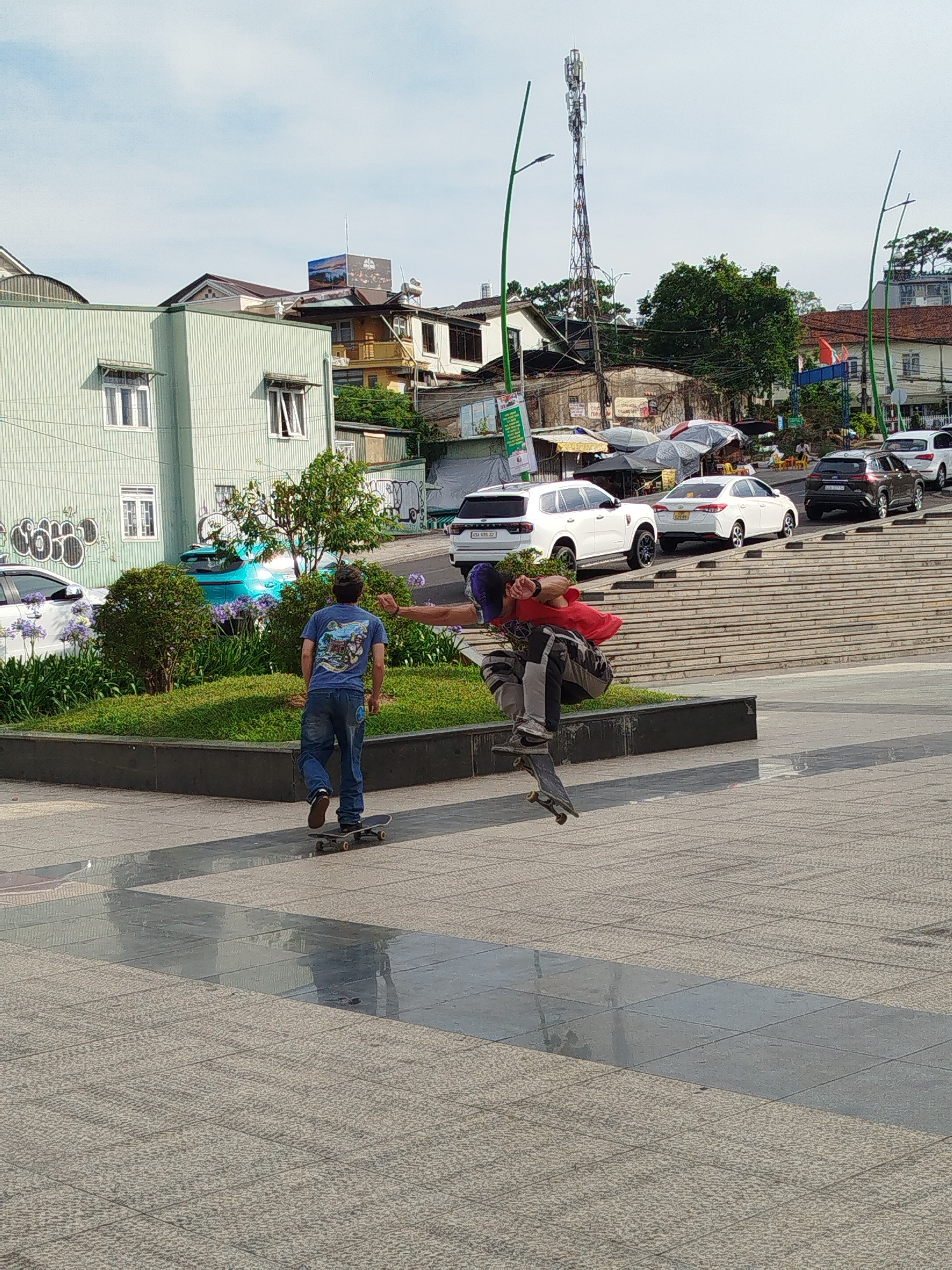 The image shows two people skateboarding in Da Lat, Vietnam. One individual, facing away from the camera, is riding on a skateboard in a forward direction. He is wearing a blue t-shirt and jeans. The second individual is airborne, performing a trick with his skateboard, wearing a red shirt, gray pants, and a purple hat.
In the background, there is a tiered concrete structure leading up to a street where several cars are parked. A green-colored building is to the left. Further in the background, the city is visible, including buildings and a tall communication tower. The sky is overcast, and there are green streetlights and vegetation in the mid-ground. The ground is paved with rectangular stones of different colors and textures.
