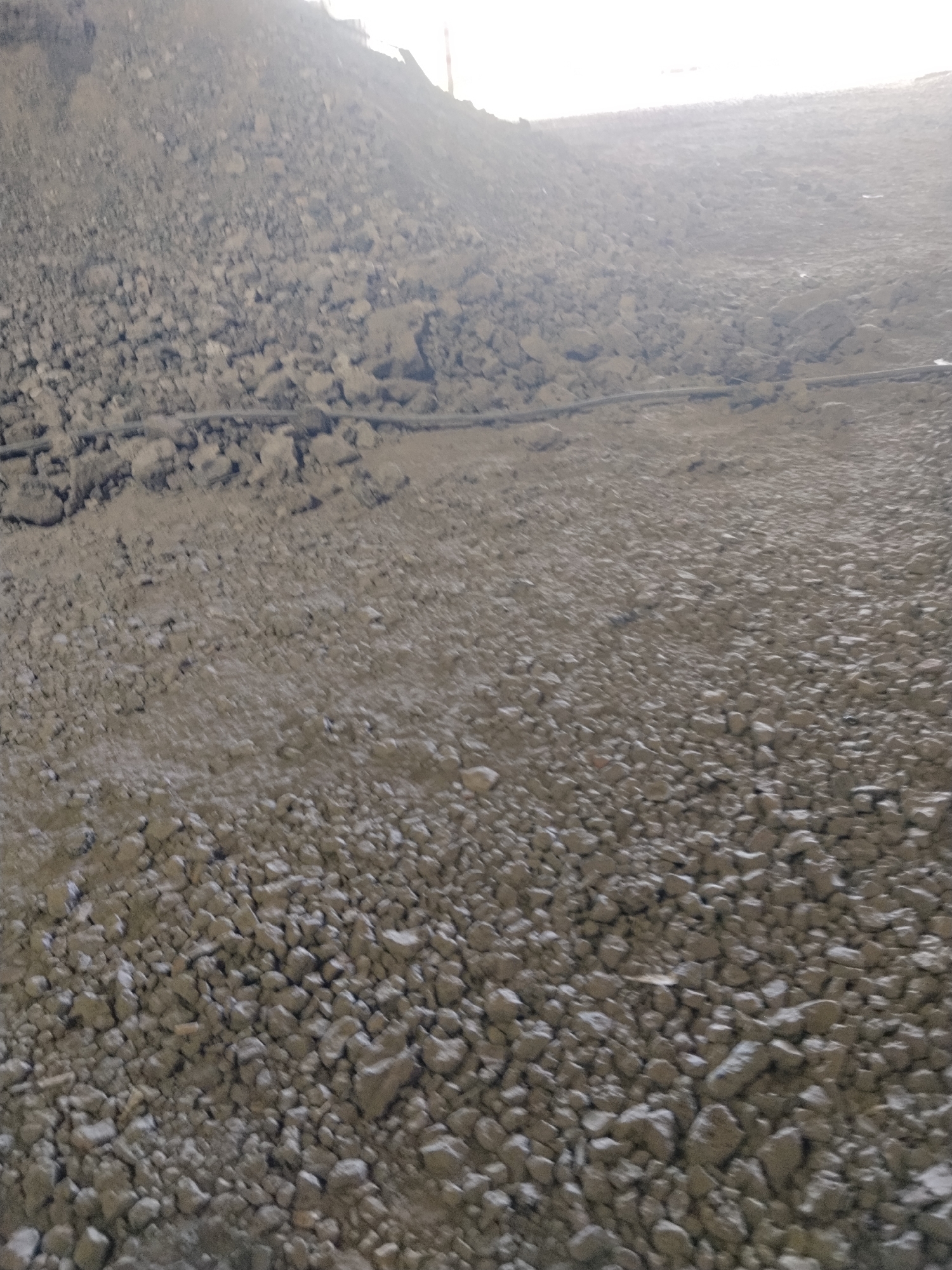 The image depicts a large, desolate landscape dominated by piles of grey, rocky debris. The terrain appears to be a construction site or an industrial area, with a significant incline in the background. A dark, horizontal line, possibly a pipe or cable, is visible traversing the middle ground of the scene. The sky overhead is bright and washed out, suggesting either daylight or artificial illumination. There are no people, animals, or vehicles visible in the frame. The overall impression is one of barrenness and industrial activity, possibly related to mining or large-scale earthmoving operations. The location is Dąbrowa Górnicza, Poland.