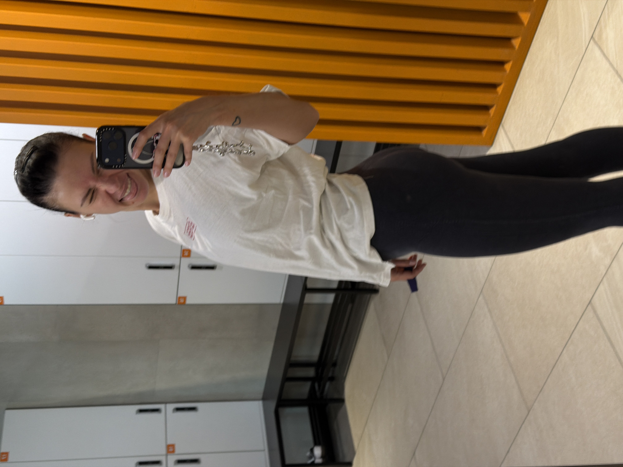 A woman is taking a mirror selfie in what appears to be a locker room or fitness center. She is wearing black leggings and a white t-shirt, and she is holding a smartphone to take the picture. The background shows white lockers with black handles, a black bench, and beige tiled flooring. An orange slat wall is visible above the lockers. The scene is likely located indoors. There is no verifiable information in the image to determine the location in Moscow, Russia.