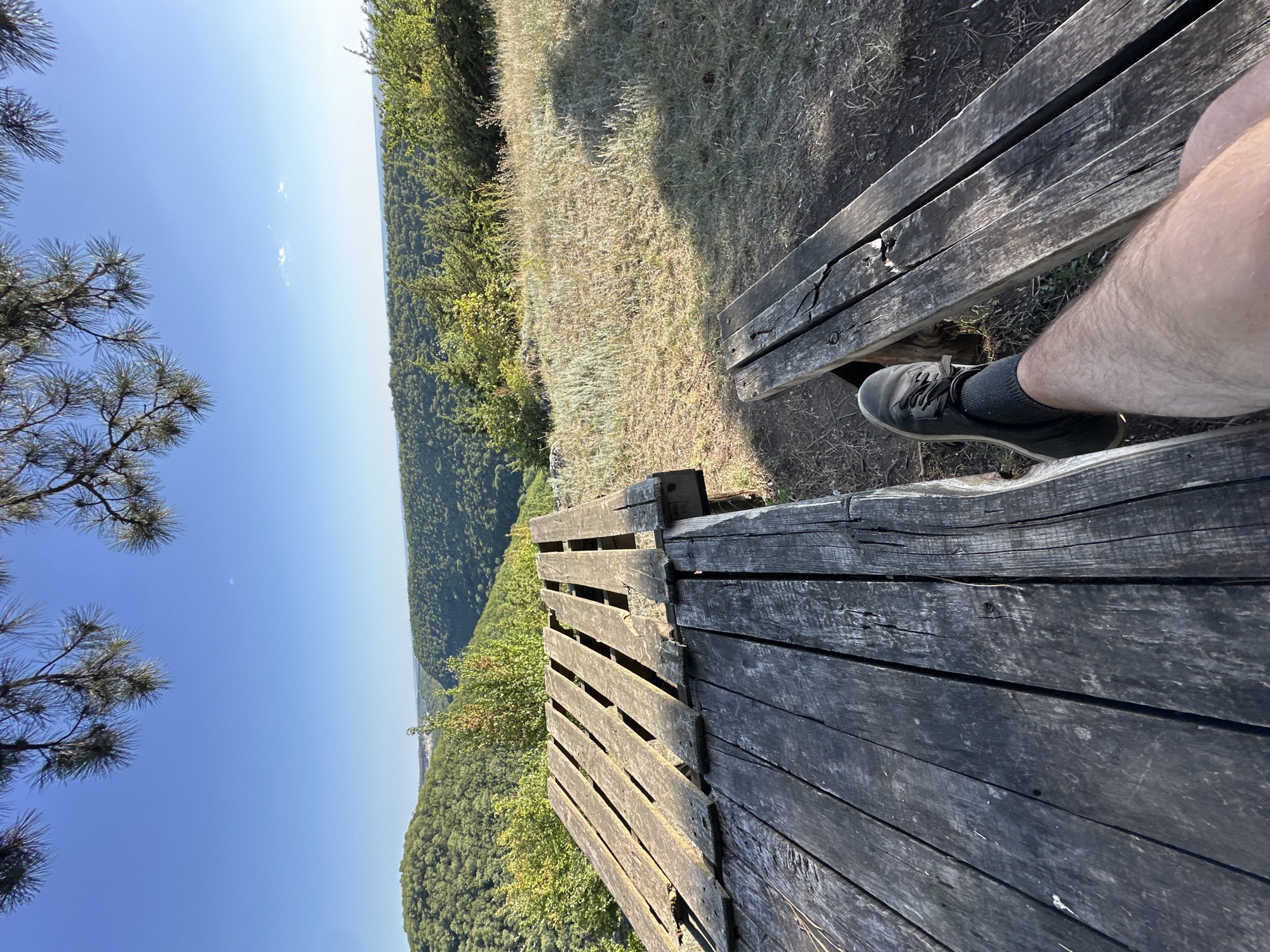 The image shows a person's leg resting on a wooden bench, with a wooden table to the left. The bench and table appear weathered and rustic. In the background, there is a lush green forest covering rolling hills, with a clear blue sky above. The scene suggests a scenic overlook or picnic area in a natural setting. The location is identified as being in the city of Makiv, Ukraine.
