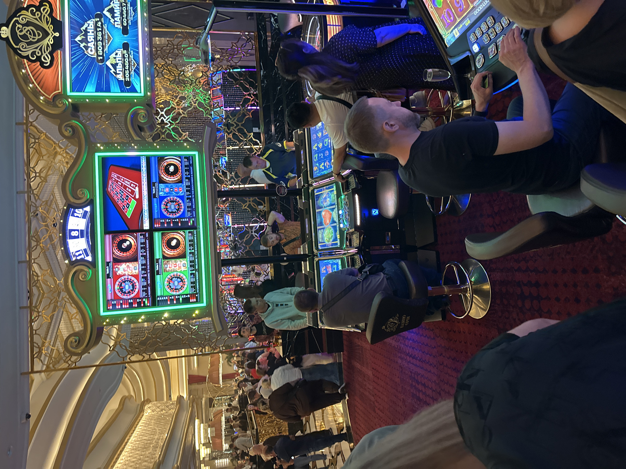 The image captures a bustling casino floor at the Sochi Casino & Resort in Estosadok, Russia. In the foreground, several patrons are engaged with slot machines. One man, wearing a black t-shirt and blue jeans, is seated and actively playing a slot machine, his hands positioned over the control panel. To his right, a woman with blonde hair is partially visible, also seemingly engaged in gaming. Another man, in a grey t-shirt, is seated at a different slot machine in the mid-ground, facing away from the camera. Behind him, a man in a blue and yellow shirt stands, observing.
The casino environment is opulent, with a large, ornate structure featuring multiple screens displaying game information, likely roulette and jackpot amounts. These screens are brightly lit and surround a central gaming area. The floor is carpeted in a deep red, with scattered gaming chairs and stools. In the background, a large crowd of people mills about, suggesting a lively atmosphere. The overall impression is one of a busy and vibrant entertainment venue.