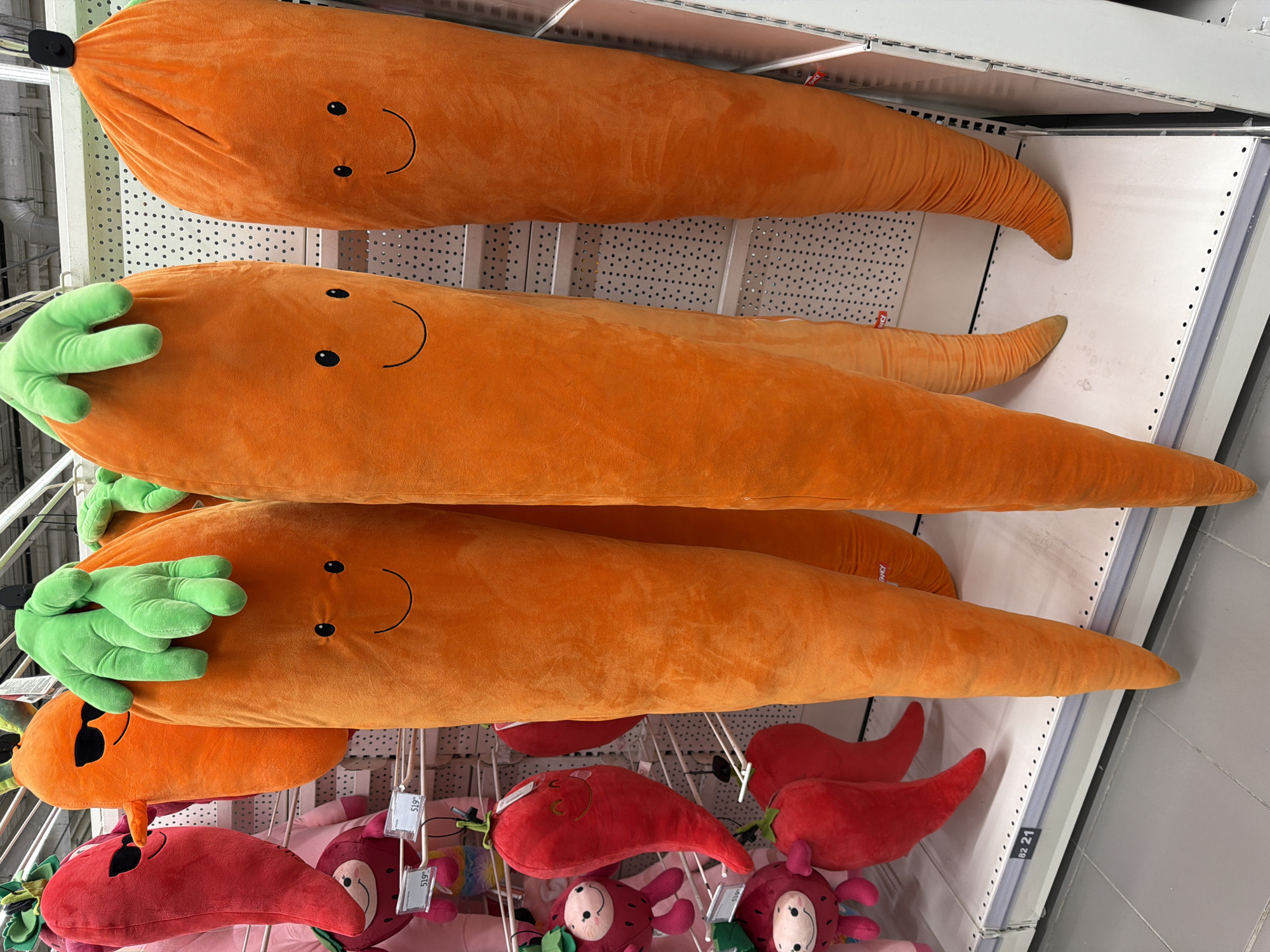 The image shows a shelf in what appears to be a retail store. The shelf is stocked with large, orange plush toys shaped like carrots, some with green plush tops. Below the carrots are several red plush toys that seem to be shaped like chili peppers or radishes, some with small faces. The store's interior is visible with white metal shelving and a light-colored floor.